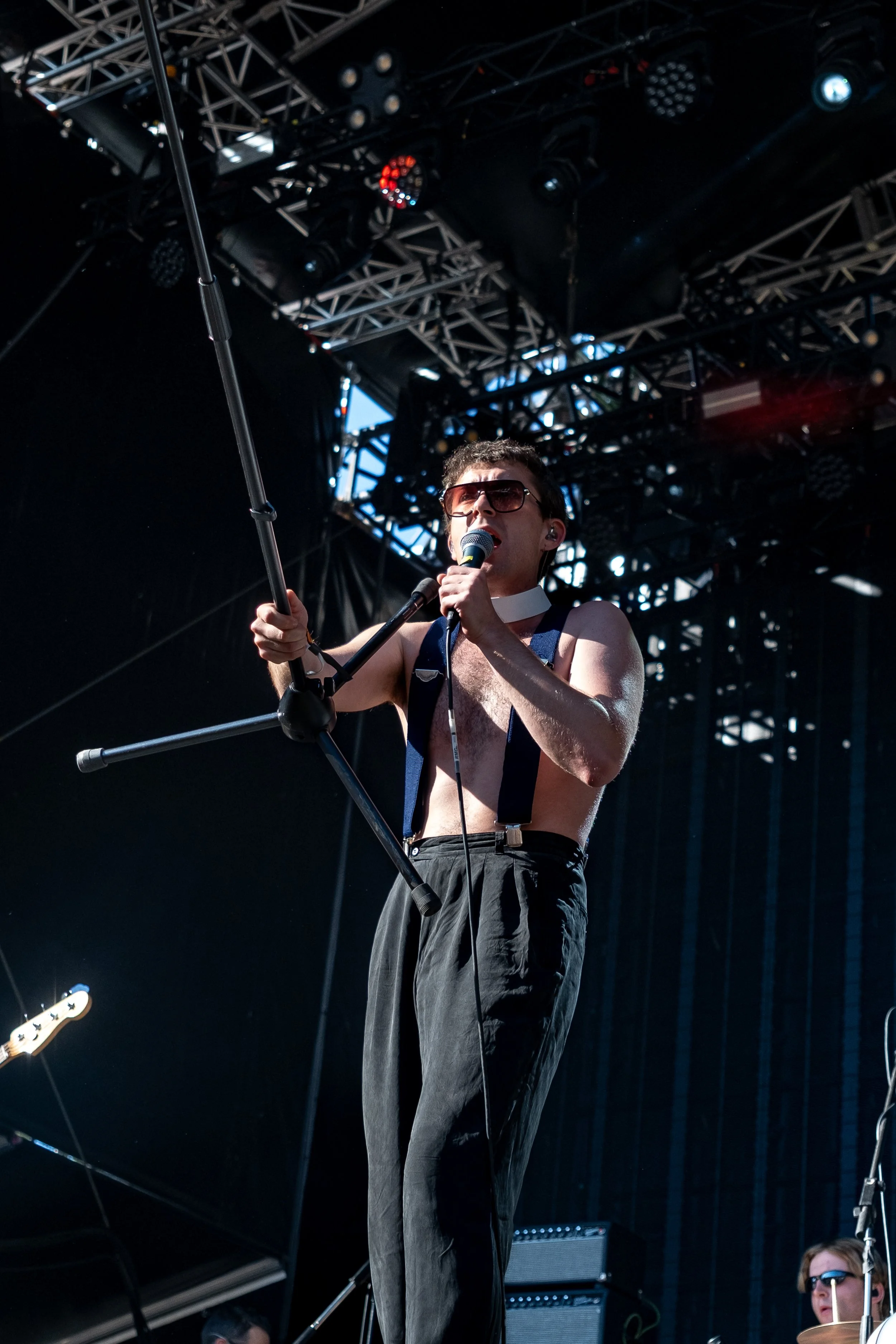 A shirtless male singer with dark hair, wearing sunglasses, suspenders, and black pants, singing into a microphone on a stage with metal scaffolding and stage lights.