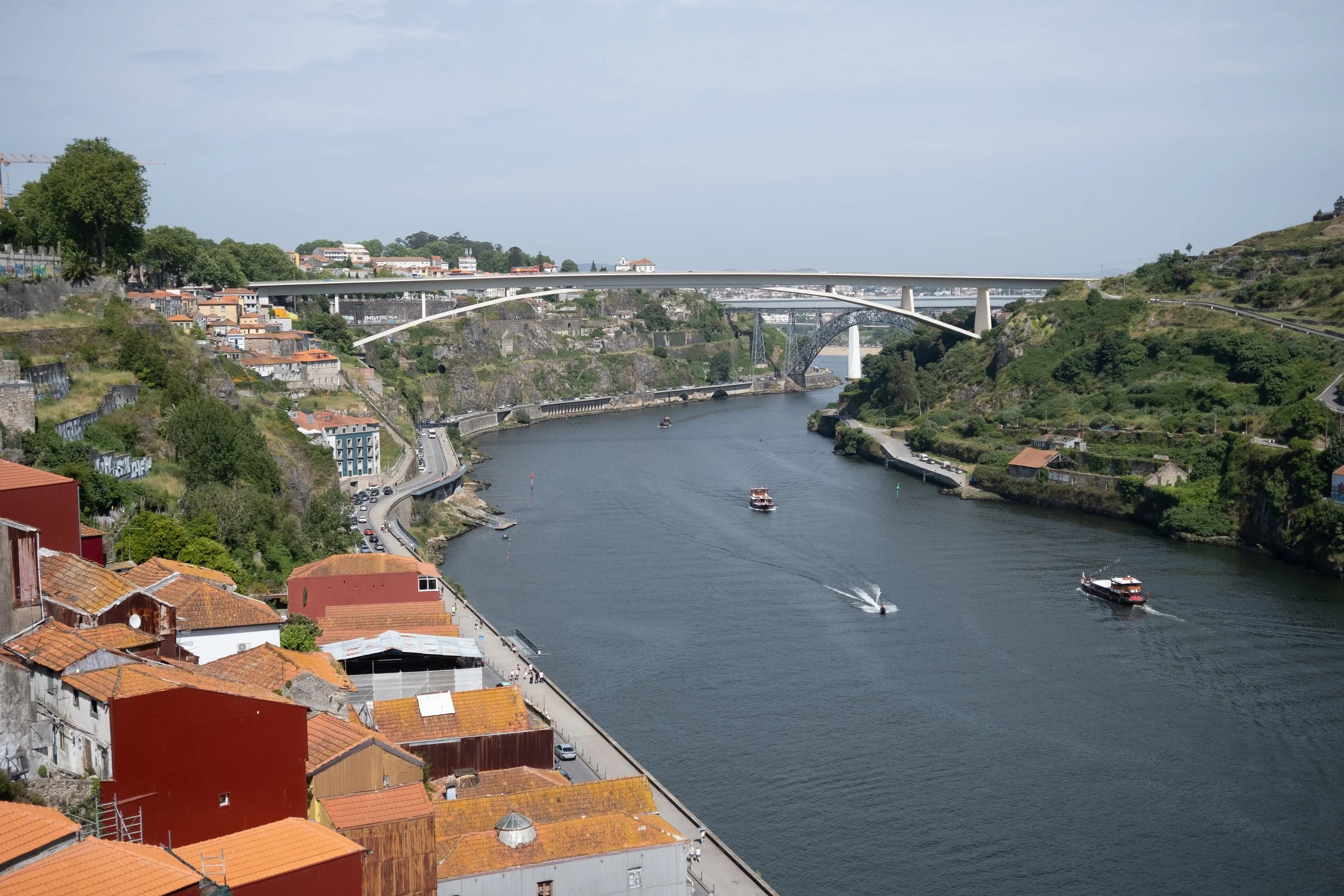 View of a river with boats, surrounded by green hills and houses, with bridges overhead in a city setting.