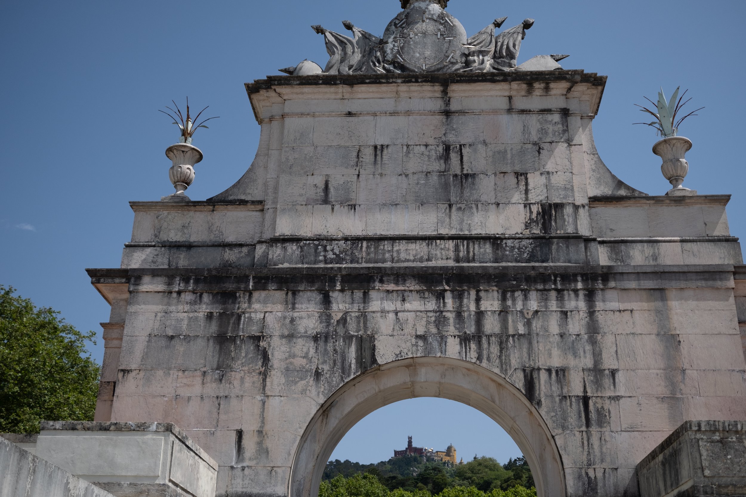 Historical stone archway with black streaks and decorative urns on top, overlooking a distant hilltop castle on a clear day.