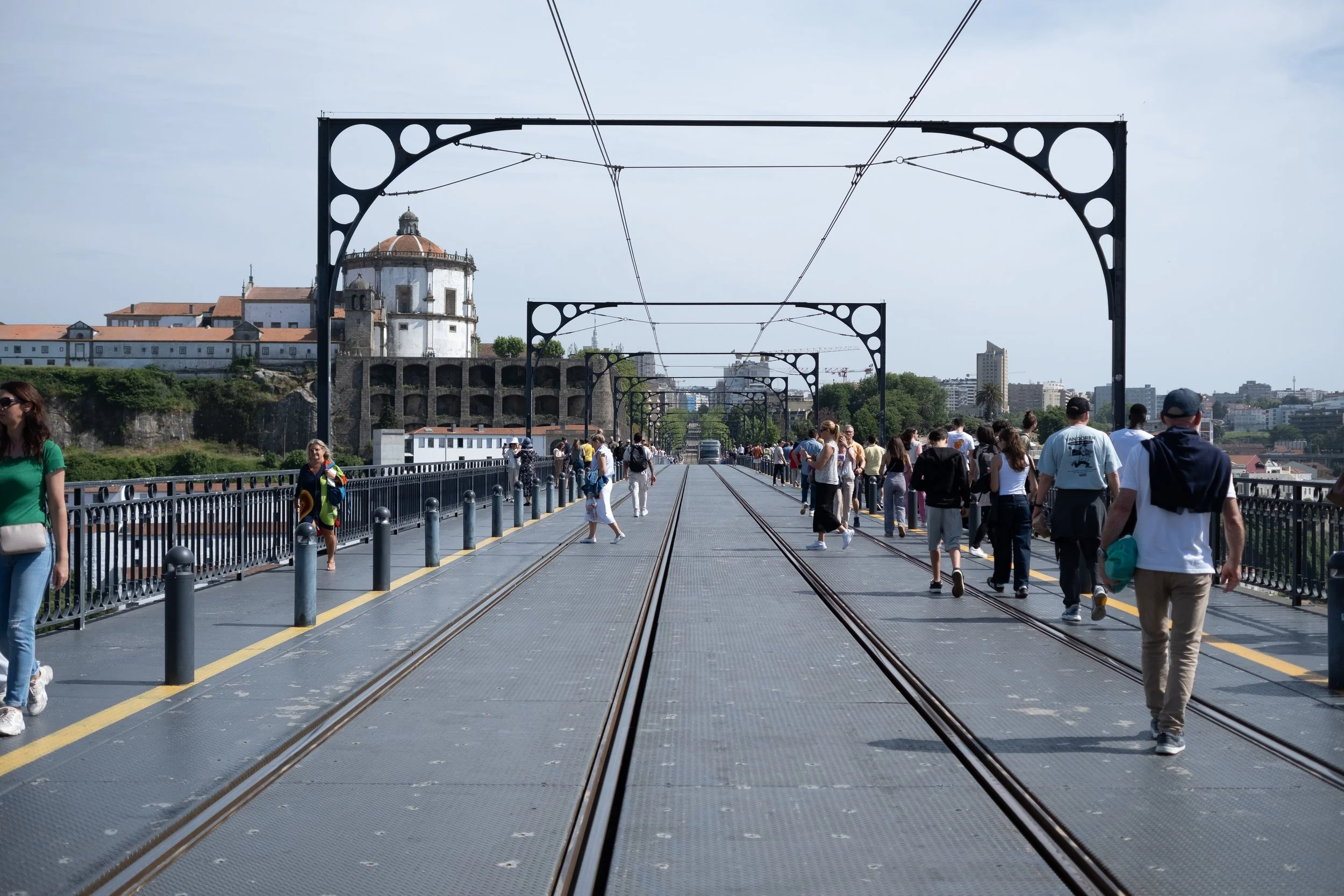 People walking on a historic bridge with tram tracks and overhead wires, overlooking city buildings and a church with a dome in the background.