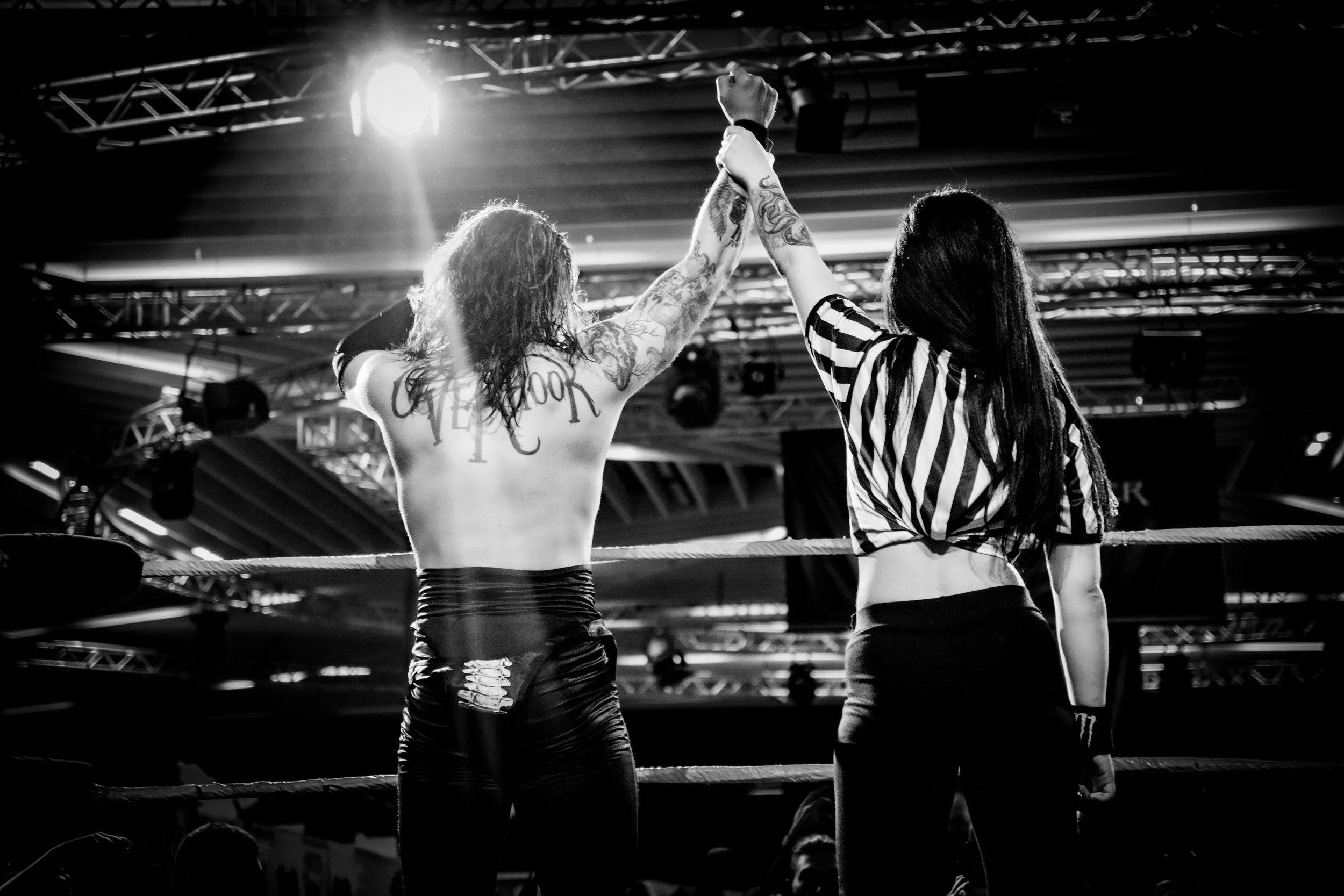 A female wrestler with tattoos on her arms raises her hand victory in a wrestling ring, while a referee with long hair and a striped shirt stands beside her in a sports arena with stage lighting and a metal truss ceiling.