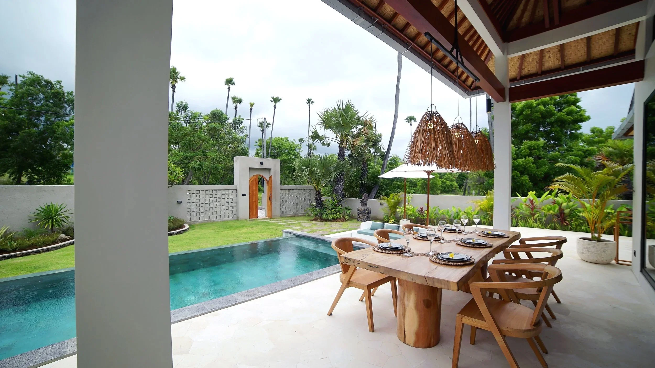 An outdoor patio with a wooden dining table set with plates and glasses, surrounded by six wooden chairs, overlooking a small pool and lush green garden with trees and plants.
