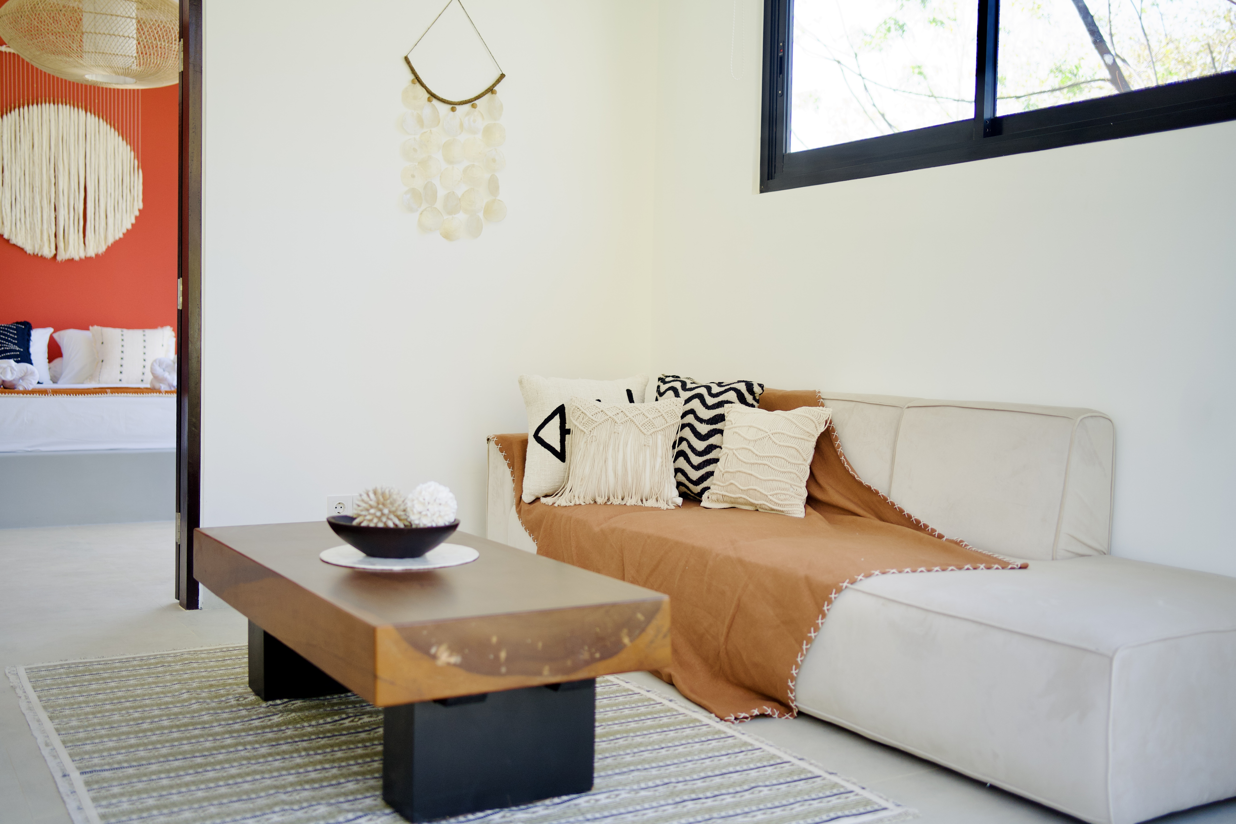 Living room with a beige sectional sofa covered with an orange throw and decorative pillows, a wooden coffee table with a black bowl and shells, a striped rug, a window with black frame, and a wall hanging made of circular shells. Part of a bedroom with white, black, and red decor is visible through an open doorway.