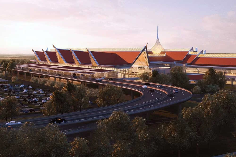 A large modern building with a distinctive roof design, surrounded by trees and a parking lot with cars, with an elevated highway in the foreground.