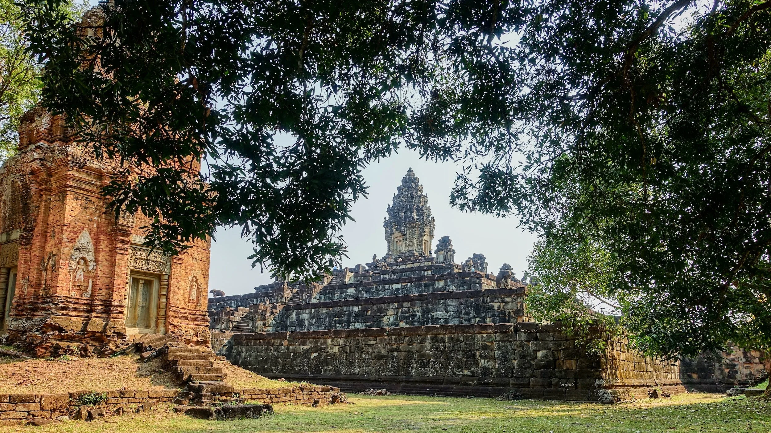 Ancient brick temple ruins with stairs and intricate carvings, framed by overhanging tree branches and leaves.