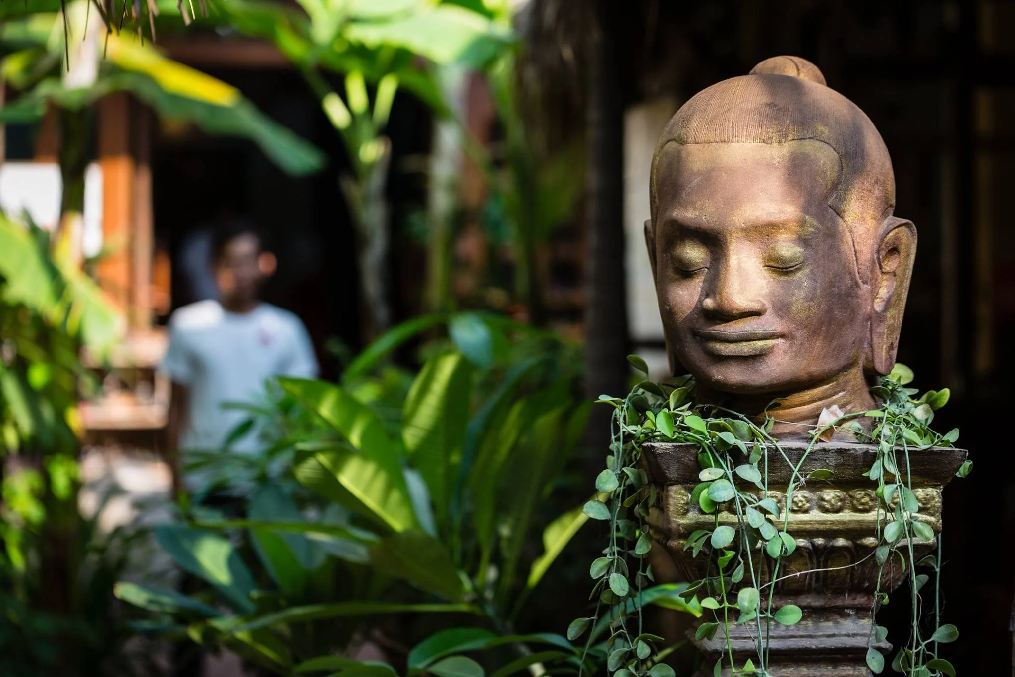 A stone sculpture of a serene face with closed eyes, surrounded by green foliage, located outdoors.