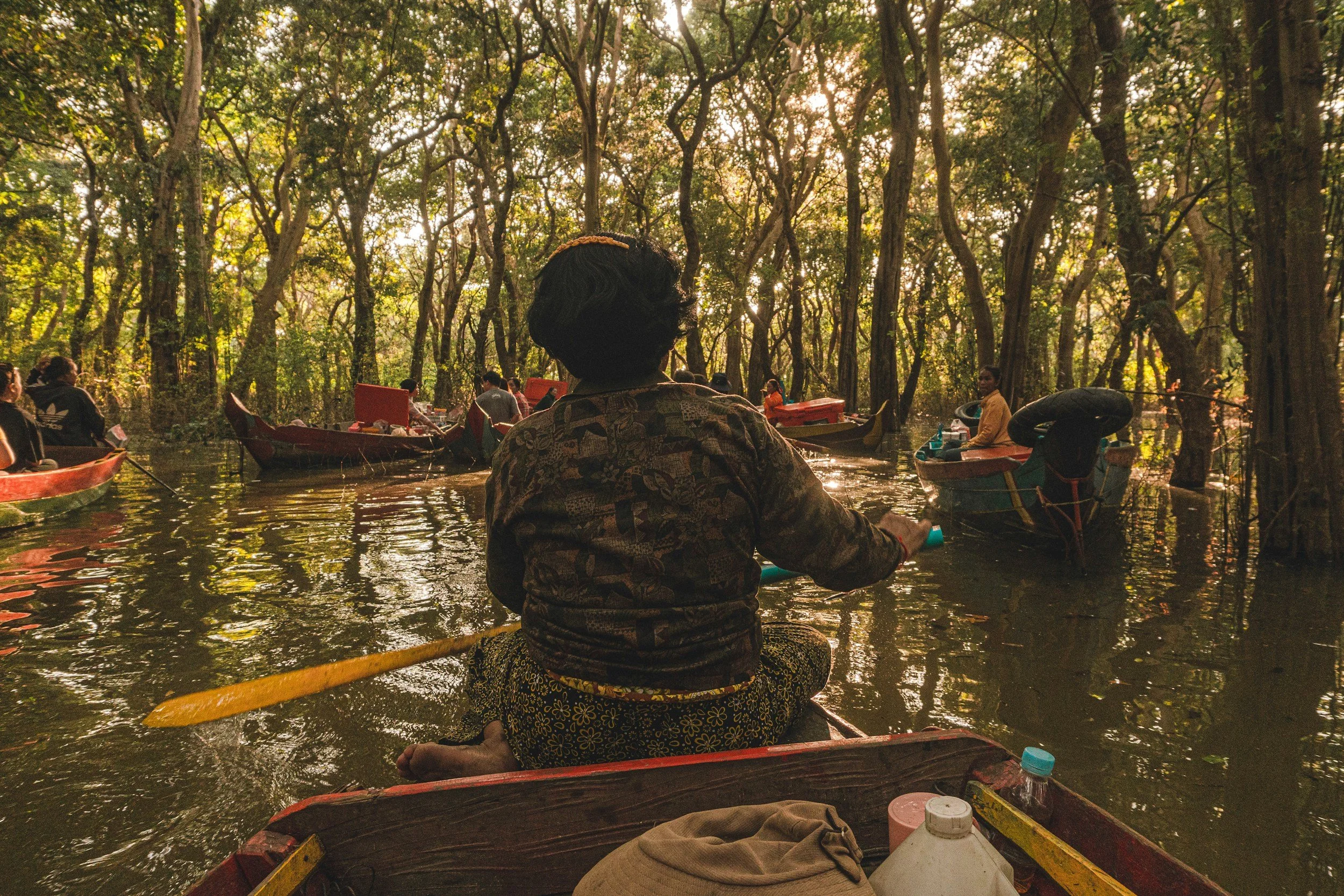 People in boats floating on a river through a forest with tall trees, sunlight filtering through the leaves, and the back of a person sitting in a boat in the foreground.