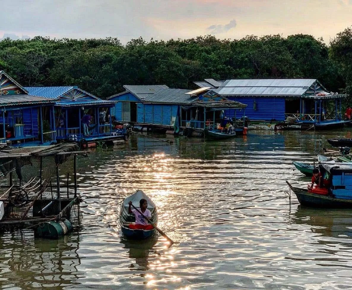 A lakeside scene with blue houses built on stilts, boats on the water, and a girl in a small boat paddling near the shore at sunset.