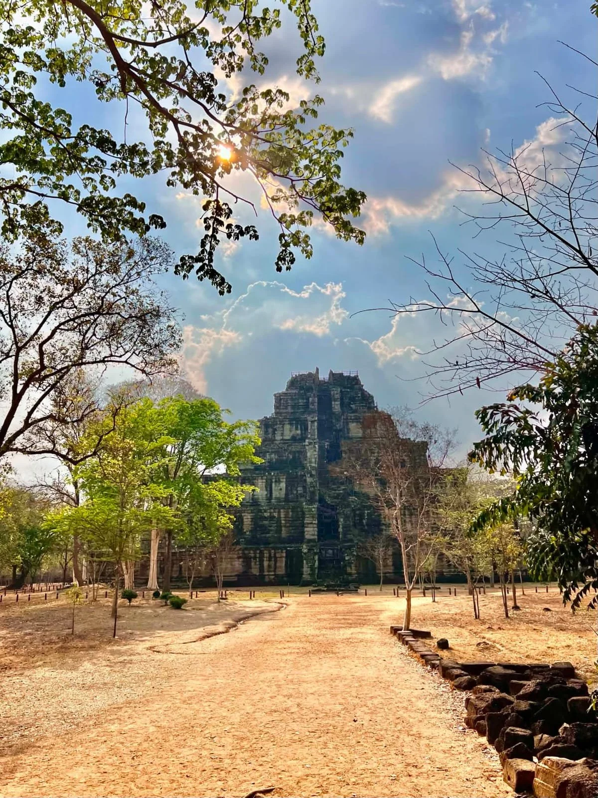 Ancient stone temple in a park with trees, dirt pathway, and a blue sky with scattered clouds.