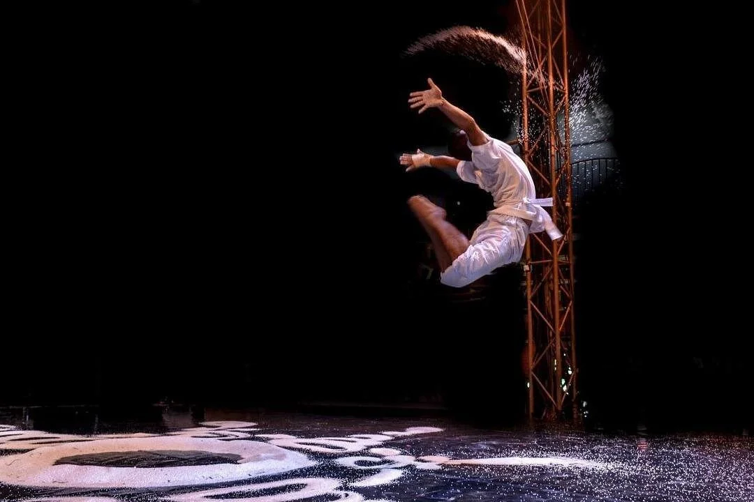 Phare Circus artist on a dark stage with rice flying around, illuminated by stage lights.