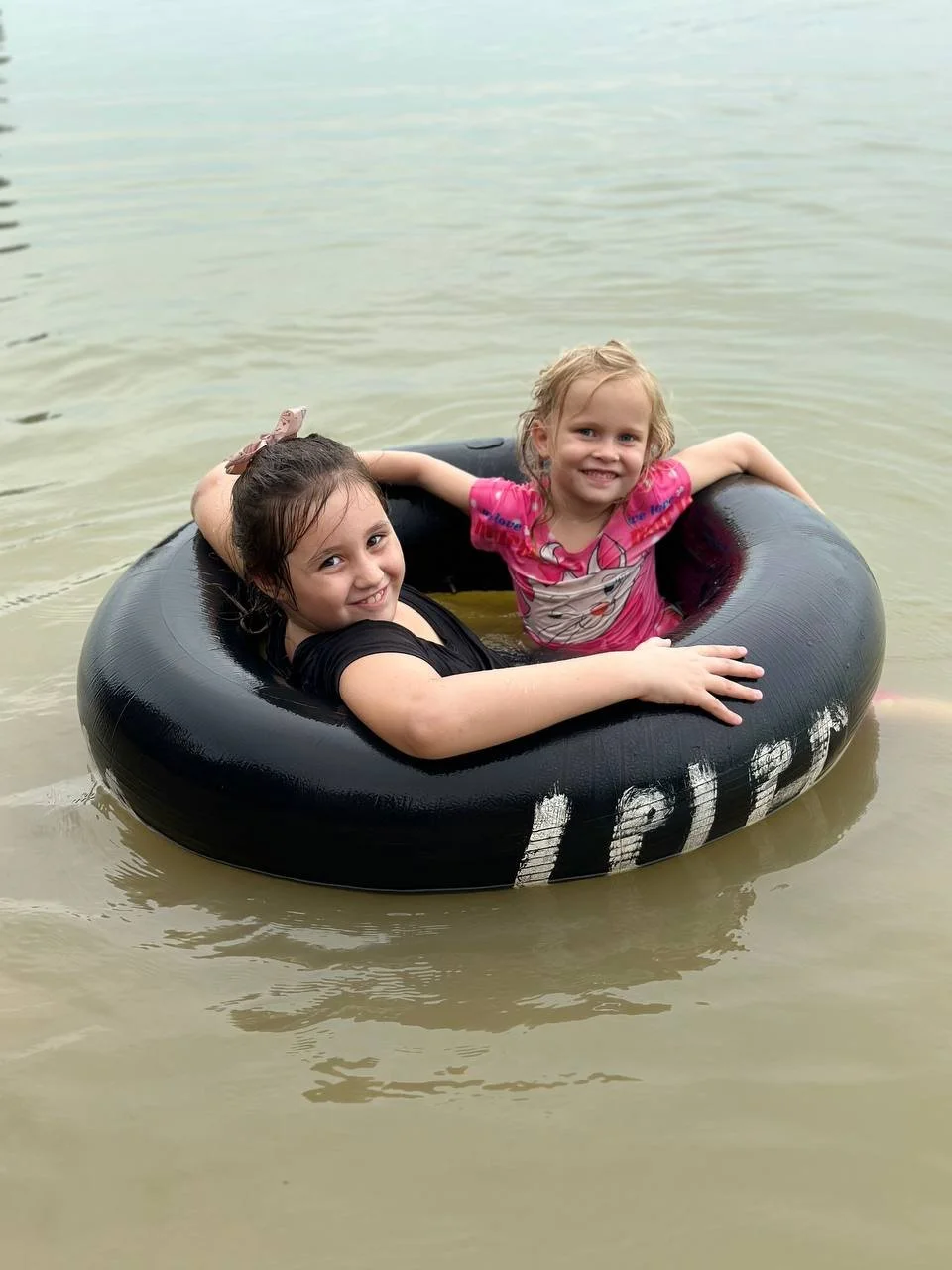 Two young girls are smiling and enjoying themselves in a black inner tube floating on a body of water.