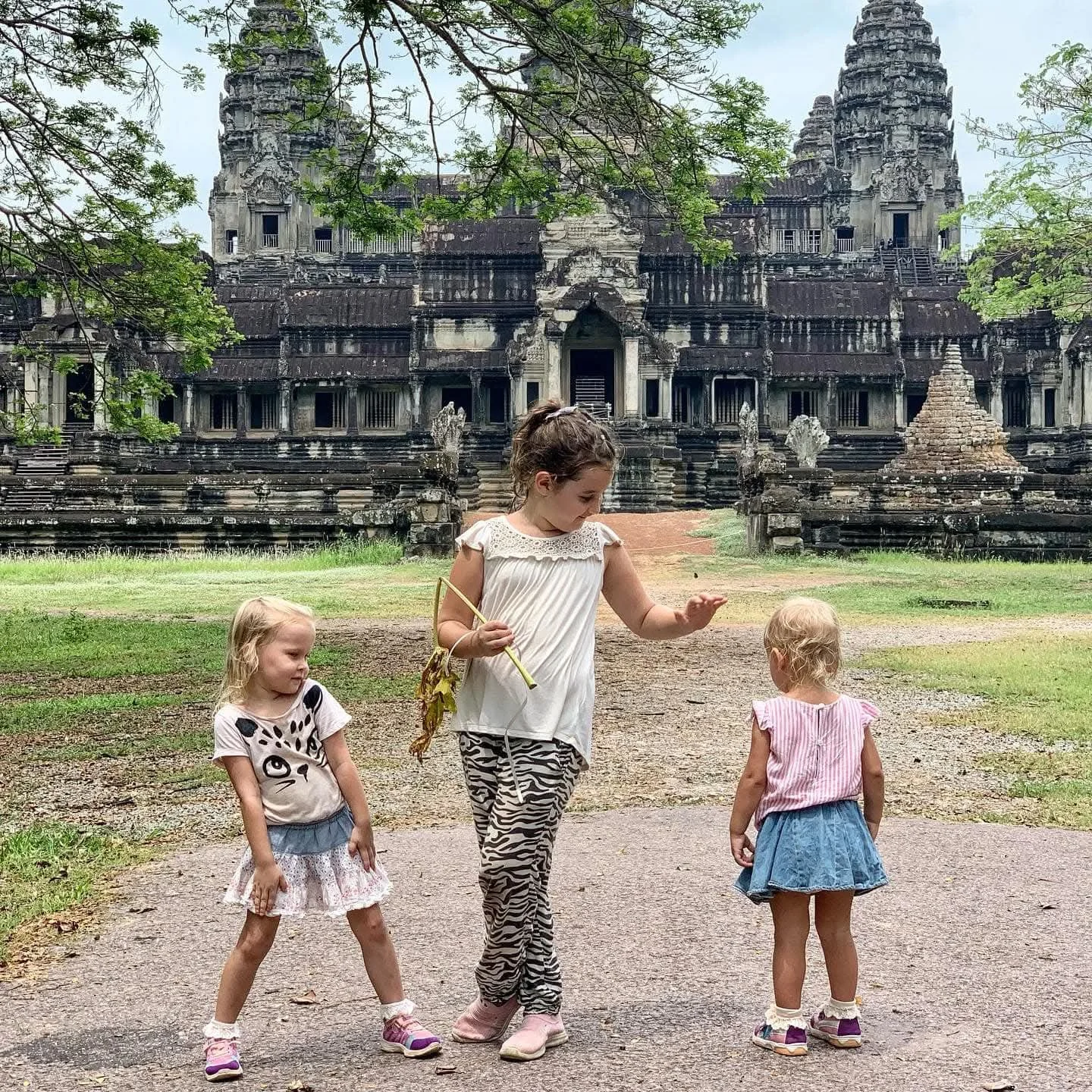 Three young girls standing on a dirt path in front of an ancient stone temple with intricate carvings, surrounded by greenery and trees.