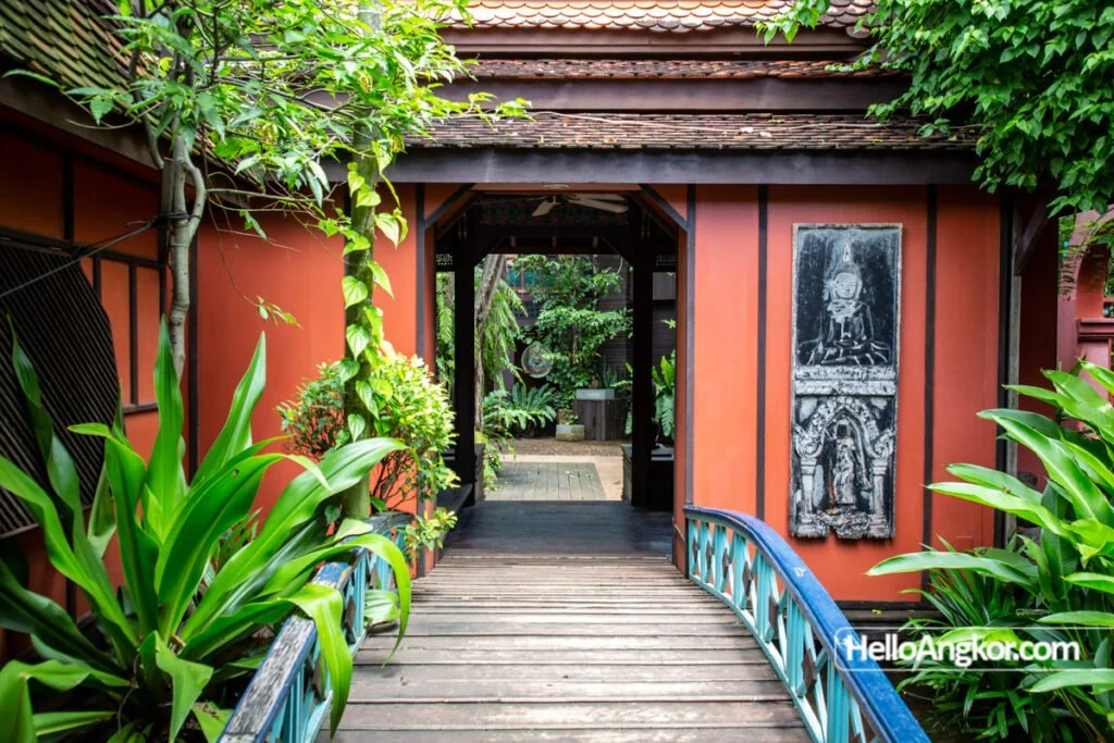 A wooden bridge leading to an entrance of a traditional building surrounded by lush green plants and trees, with an Asian-inspired decor, including a statue on the wall.