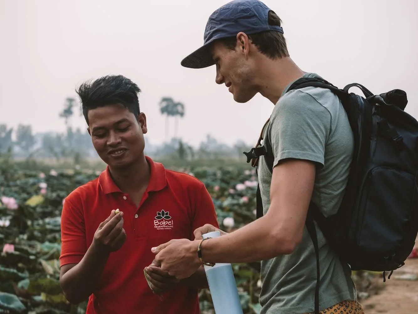 Two men stand outdoors, one wearing a red shirt with logo, the other wearing a gray shirt, a backpack, and a blue cap. They are looking at an object in the man's hand, with a field of plants and some palm trees in the background.