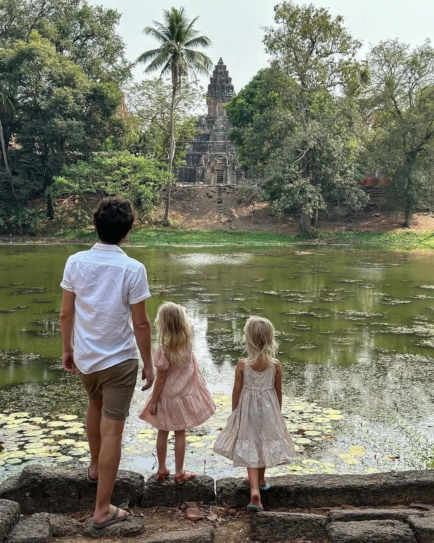 A man with three young girls standing at the edge of a pond, looking at a temple ruins in the distance surrounded by trees and lush greenery.