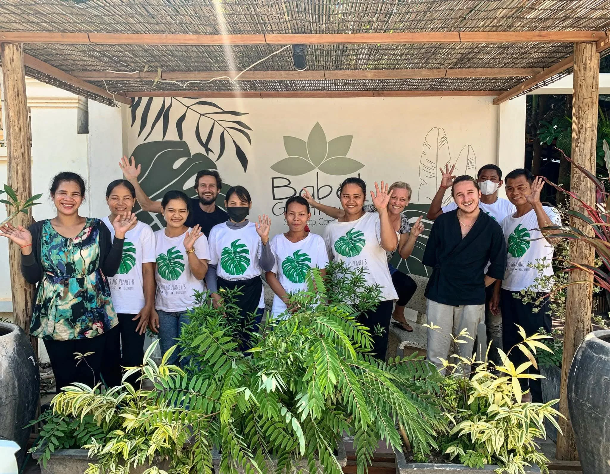 Group of people standing outdoors behind a variety of green plants, waving and smiling, with a decorative wall featuring large plant illustrations in the background.