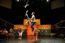 Circus performers on stage, one balancing on a tire while reading from a book, with an acrobat and other performers in the background.