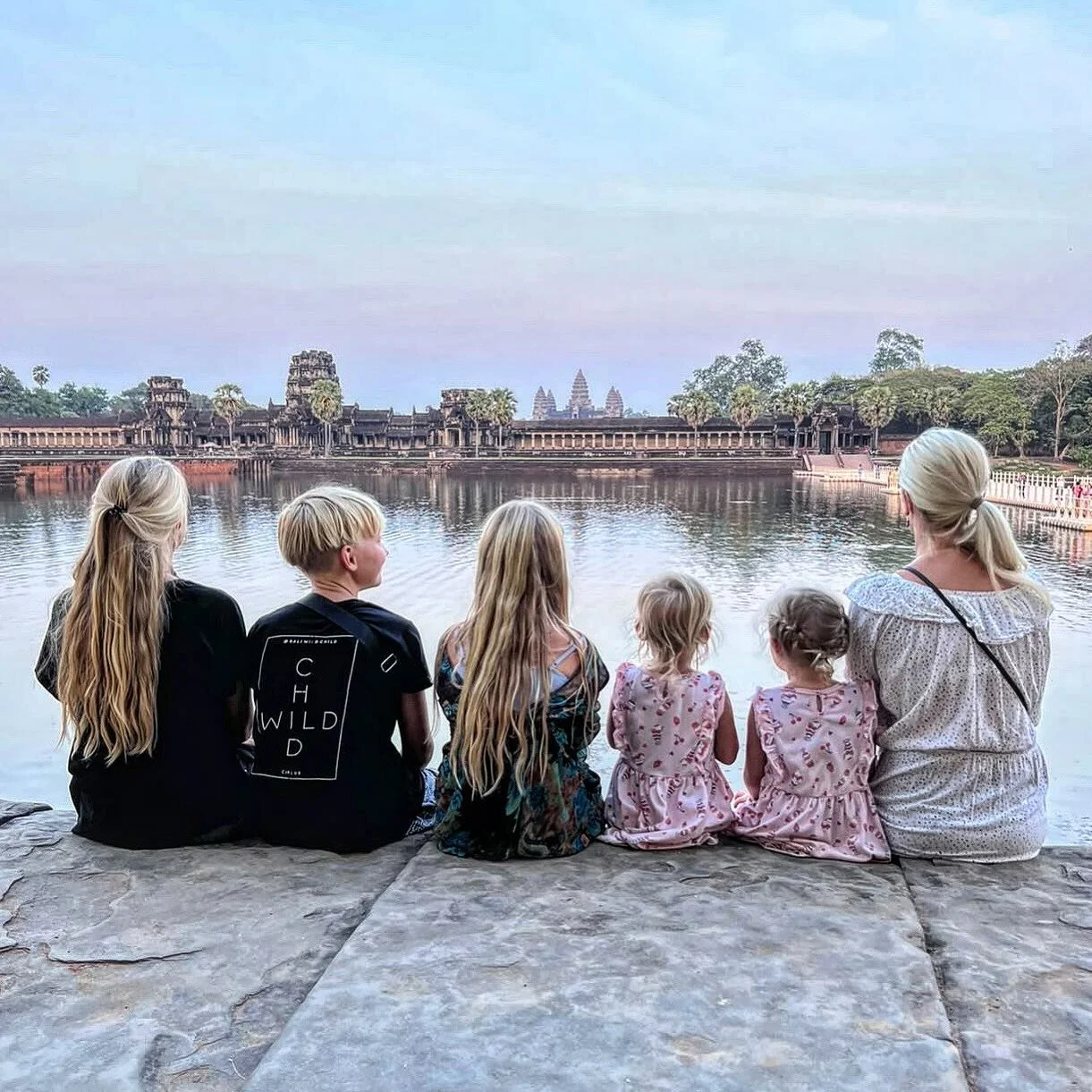Six people, including children and adults, sitting on a stone ledge by a body of water, watching a historic temple complex across the water at sunrise or sunset.