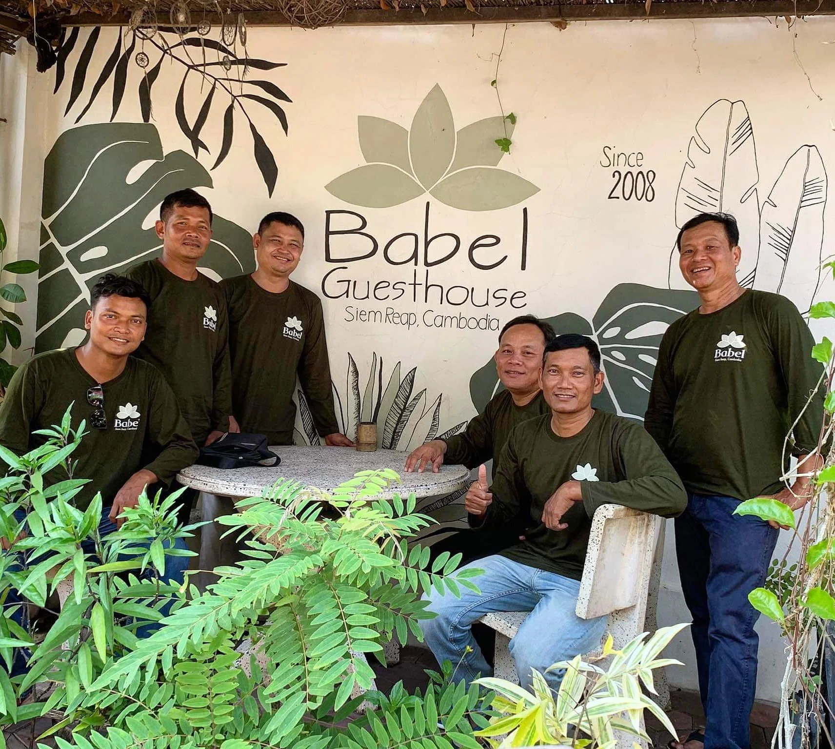 Group of six men wearing matching dark green shirts with 'Babel' logo, posing for a photo at Babel Guesthouse in Siem Reap, Cambodia, with jungle-themed wall art behind them and lush green foliage in the foreground.