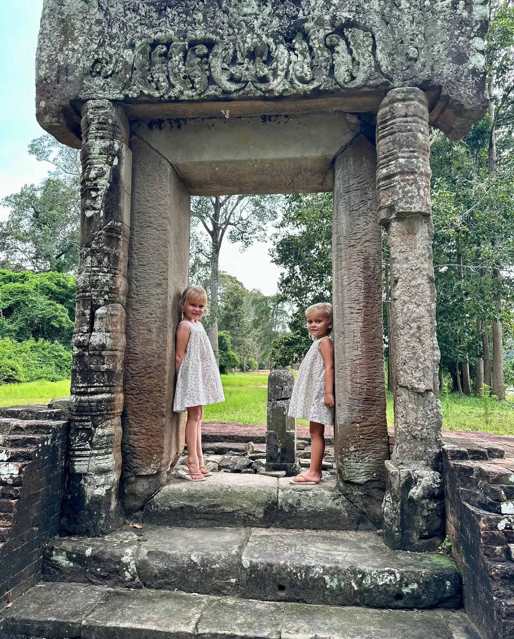 Two young girls in white dresses standing on stone platform inside ancient stone temple arch with carved decorative details, surrounded by greenery.