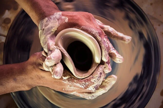 Two hands are shaping a piece of clay into a rounded form over a spinning pottery wheel, with the wheel and clay in motion.