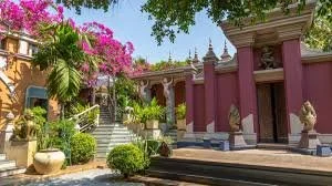A colorful courtyard with pink walls, statues, potted plants, stairs, and trees under a blue sky. Theams House, Siem Reap