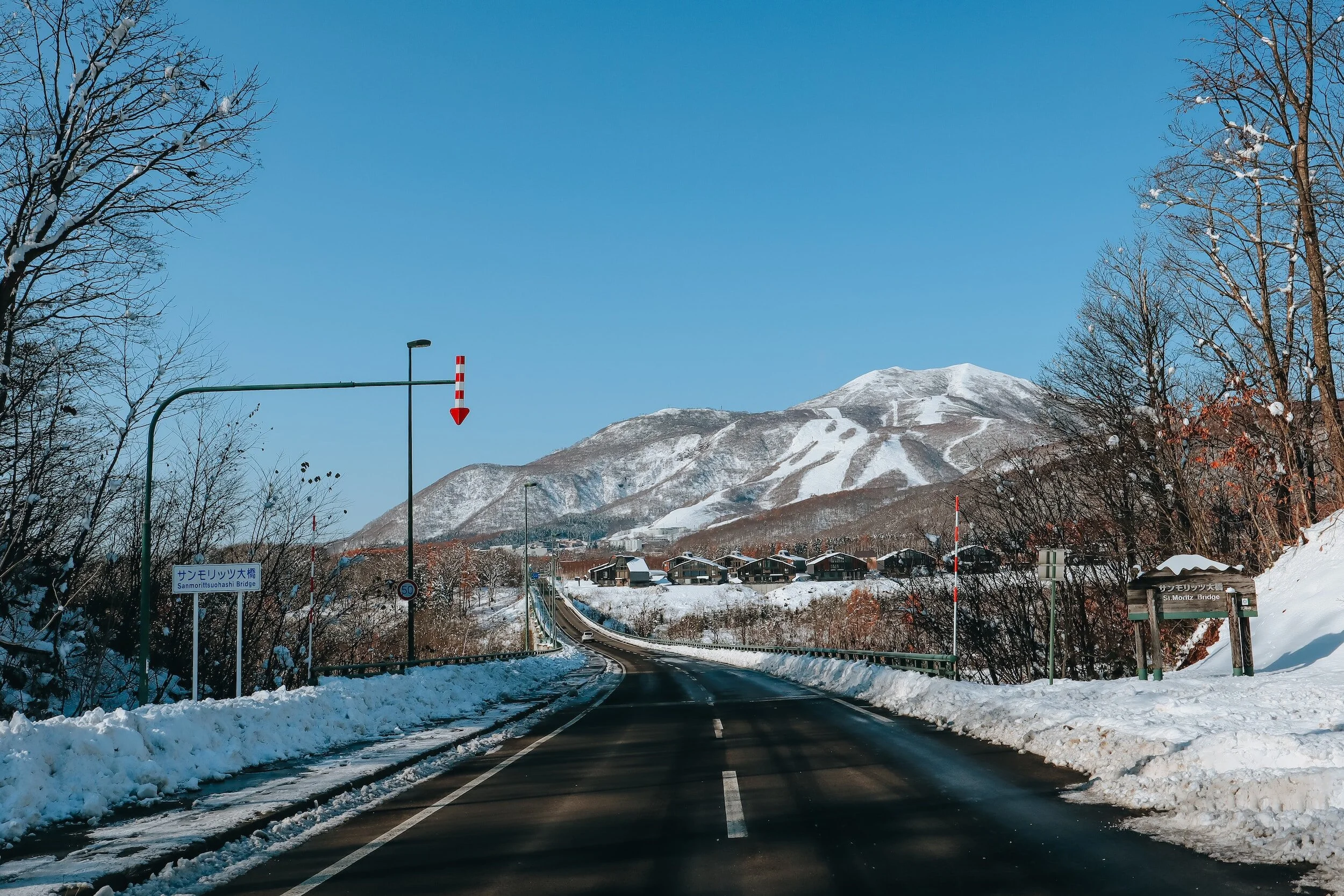 Snow-covered mountain in the background with clear blue sky, leading to a winding road lined with snow on both sides, trees, and a small bridge sign on the right.