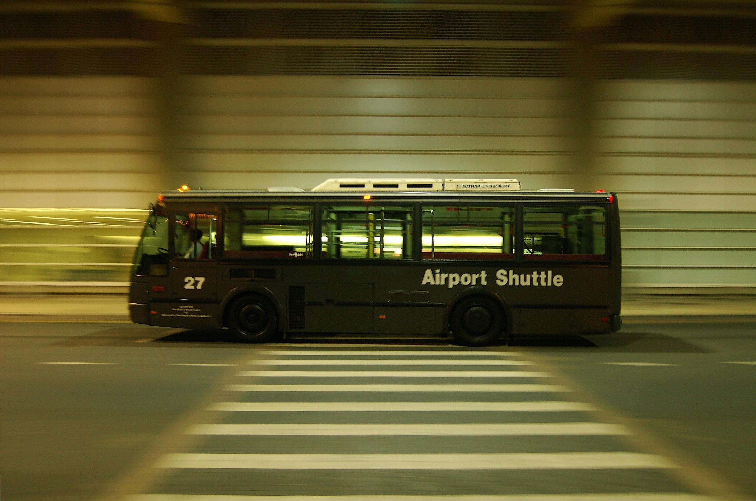 Black airport shuttle bus moving through an airport at night with blurred background and illuminated inside.
