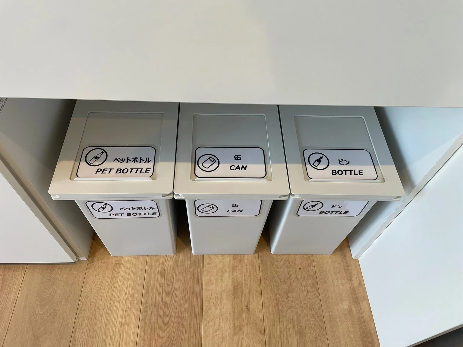 Three gray trash bins with labels for PET bottles, cans, and bottles in Japanese and English in a row on a wooden floor.