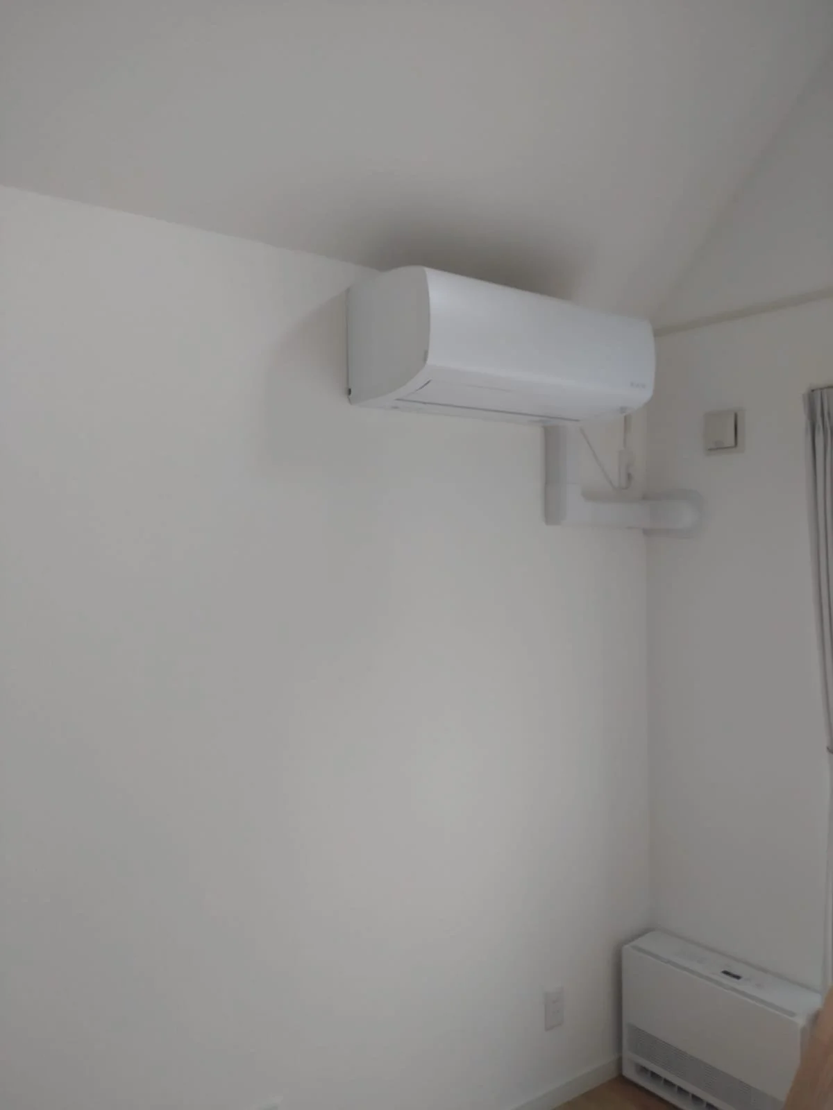 White wall-mounted air conditioner and a vertical white heater in a room with white walls and a corner window with closed curtains.