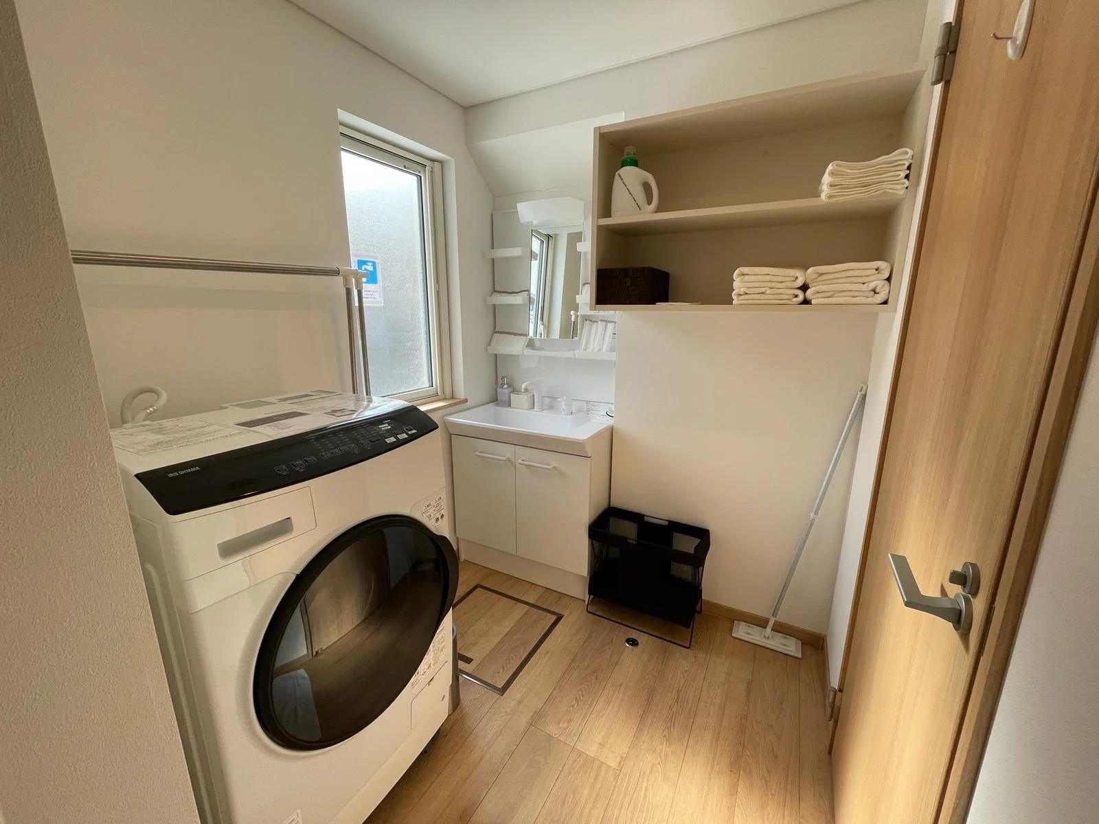A laundry room with a front-loading washing machine, a small white sink with a mirror above it, shelves with folded towels and a laundry detergent bottle, a window, a mop and a black storage rack.