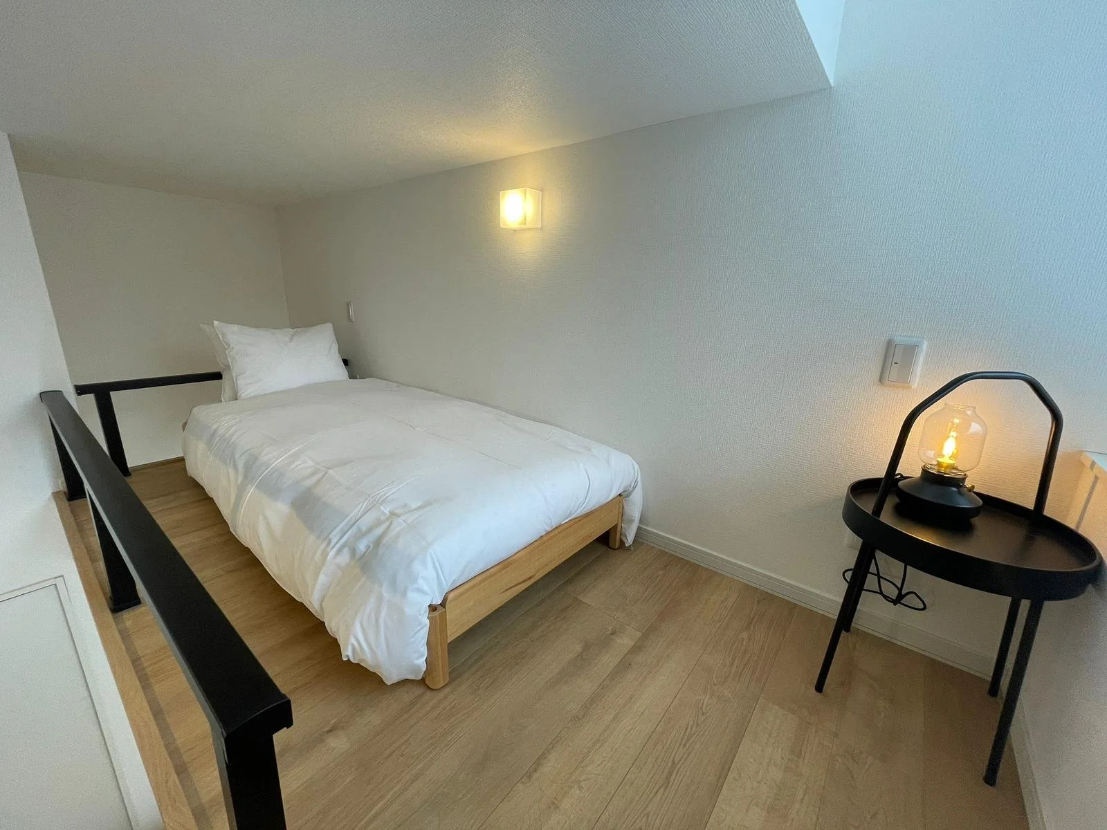 A minimalist bedroom with a white bed and pillow, black wooden railing, black side table with a lantern, beige wooden floor, white textured walls, and two wall-mounted light fixtures.