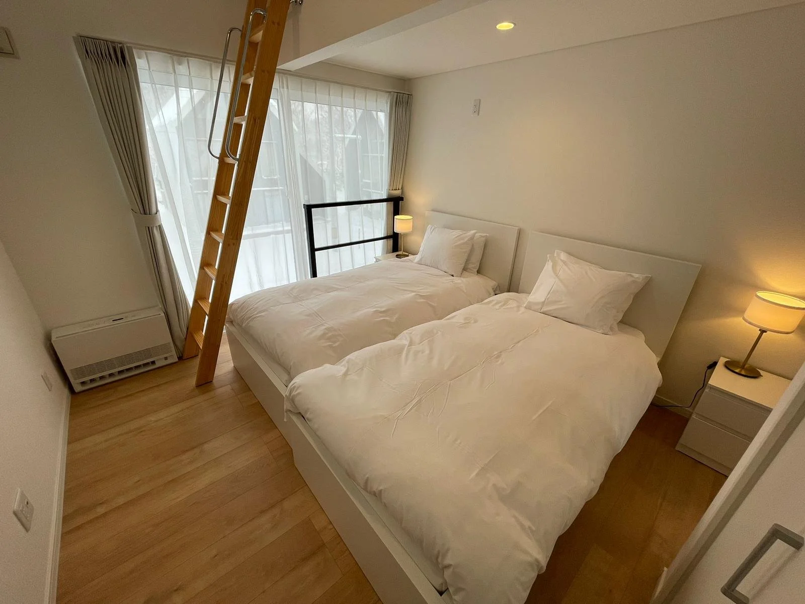 A bedroom with two single beds pushed together, each with white bedding and pillows. A small nightstand with a lamp is between the beds. There are large windows with sheer curtains and a French door leading outside. A wooden ladder leads up to a loft or elevated space.