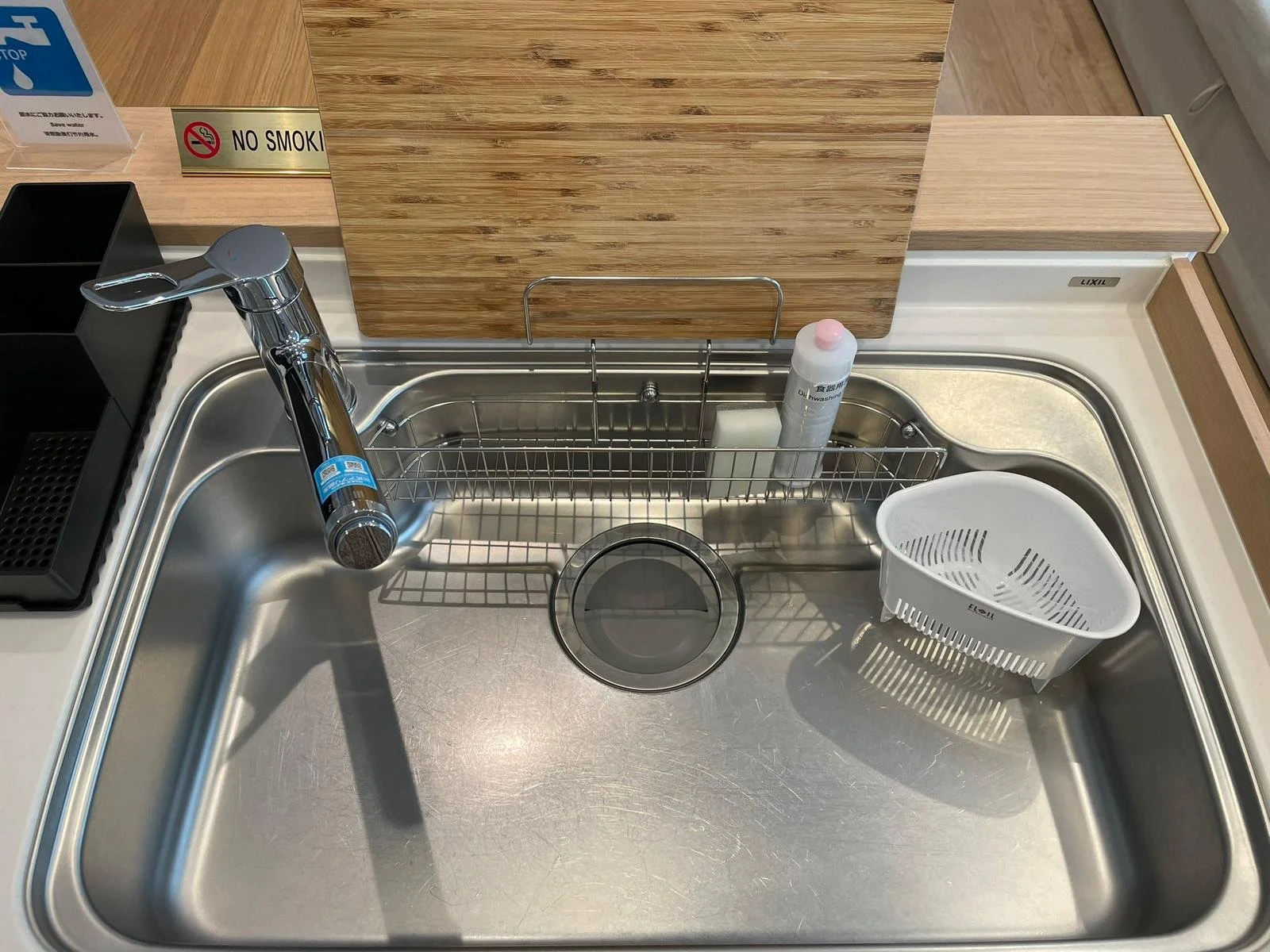 Kitchen stainless steel sink with a faucet, a white plastic colander, a soap dispenser, and a metal rack in the sink. There are wooden panels and signs nearby.
