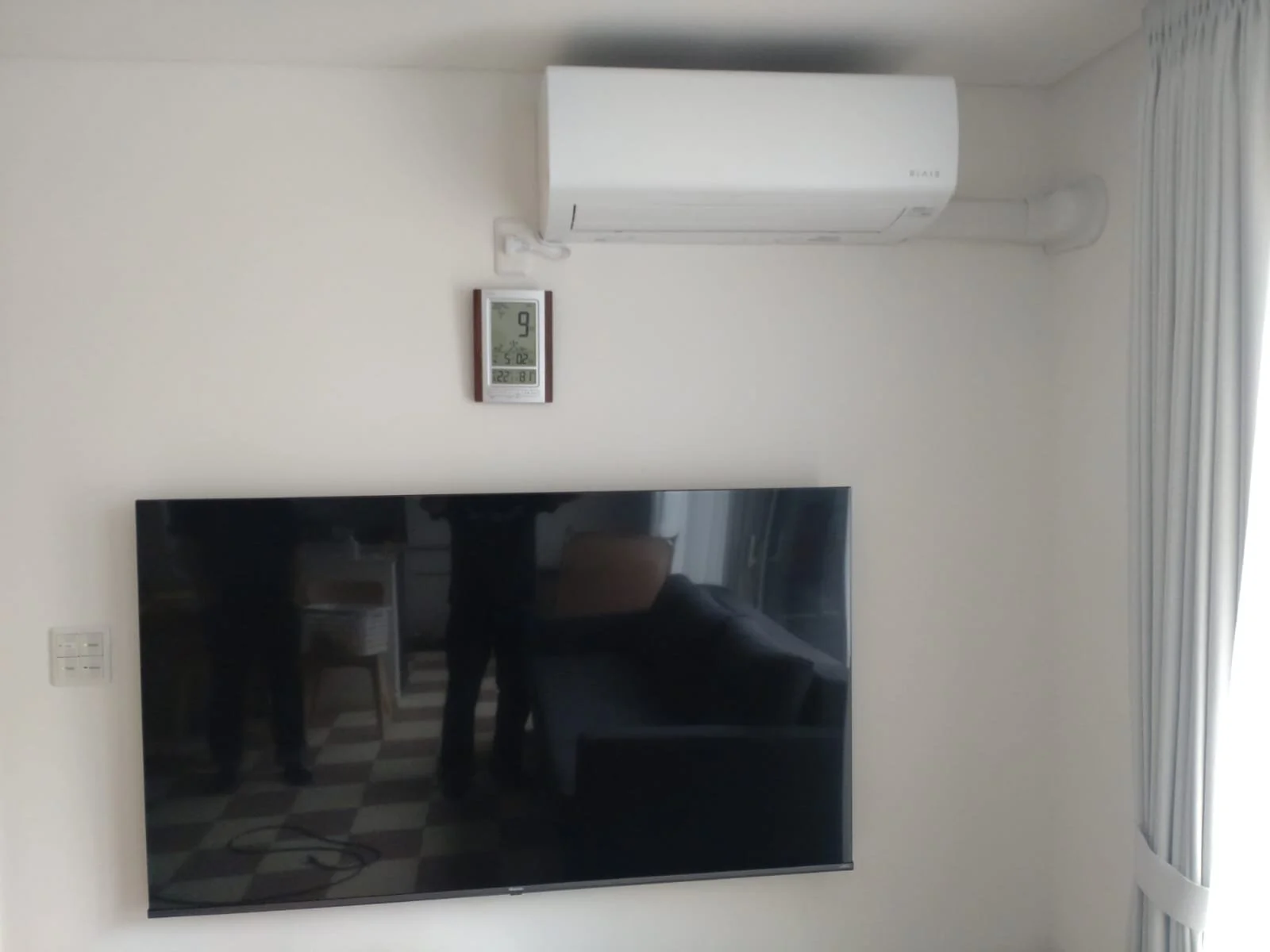 Wall-mounted flat-screen television, air conditioning unit, and a digital thermostat on a plain white wall in a modern interior room.