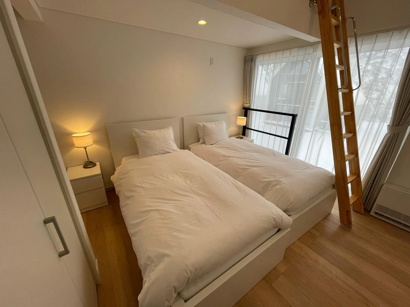 A bedroom with two twin beds, each with white bedding and pillows, beside a window with sheer curtains and a black safety rail. A wooden ladder leans against the window, and there are bedside lamps on white nightstands.