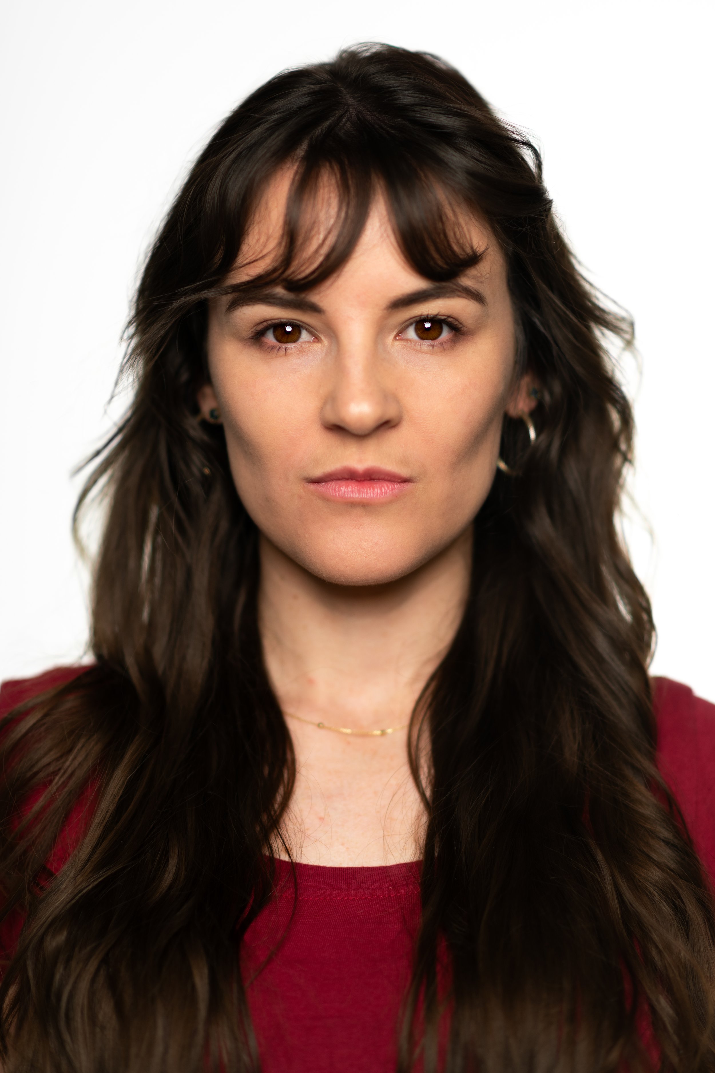 A woman with long wavy dark brown hair, wearing a dark red shirt, looking directly at the camera against a plain white background.