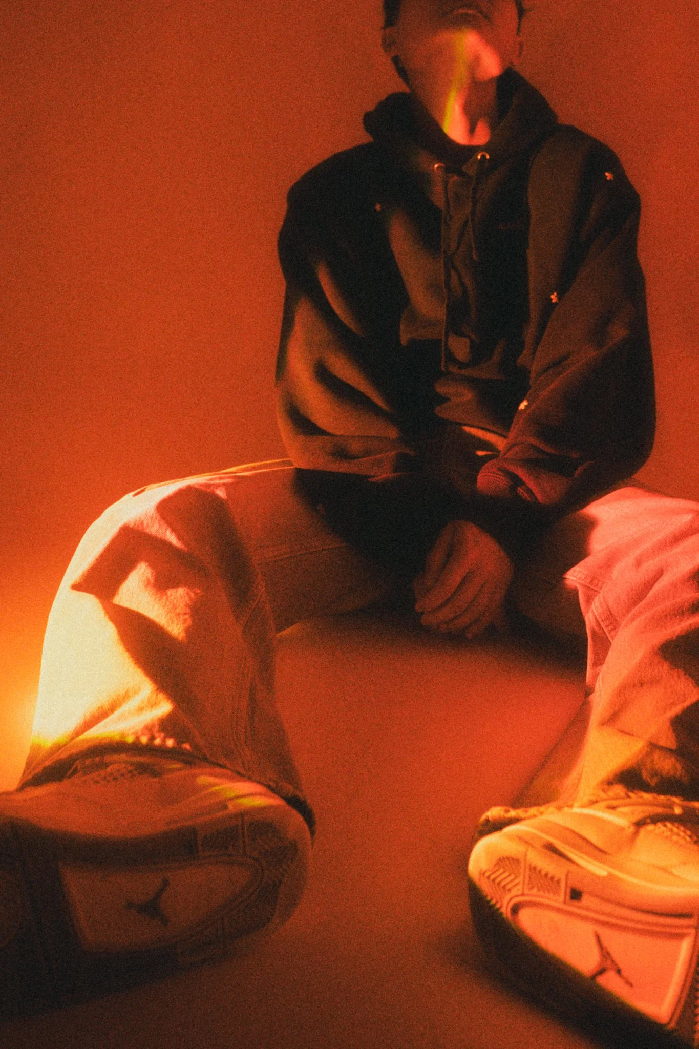 Person sitting on a bed wearing a black hoodie, with feet in athletic shoes, in a dimly lit room with warm, orange lighting