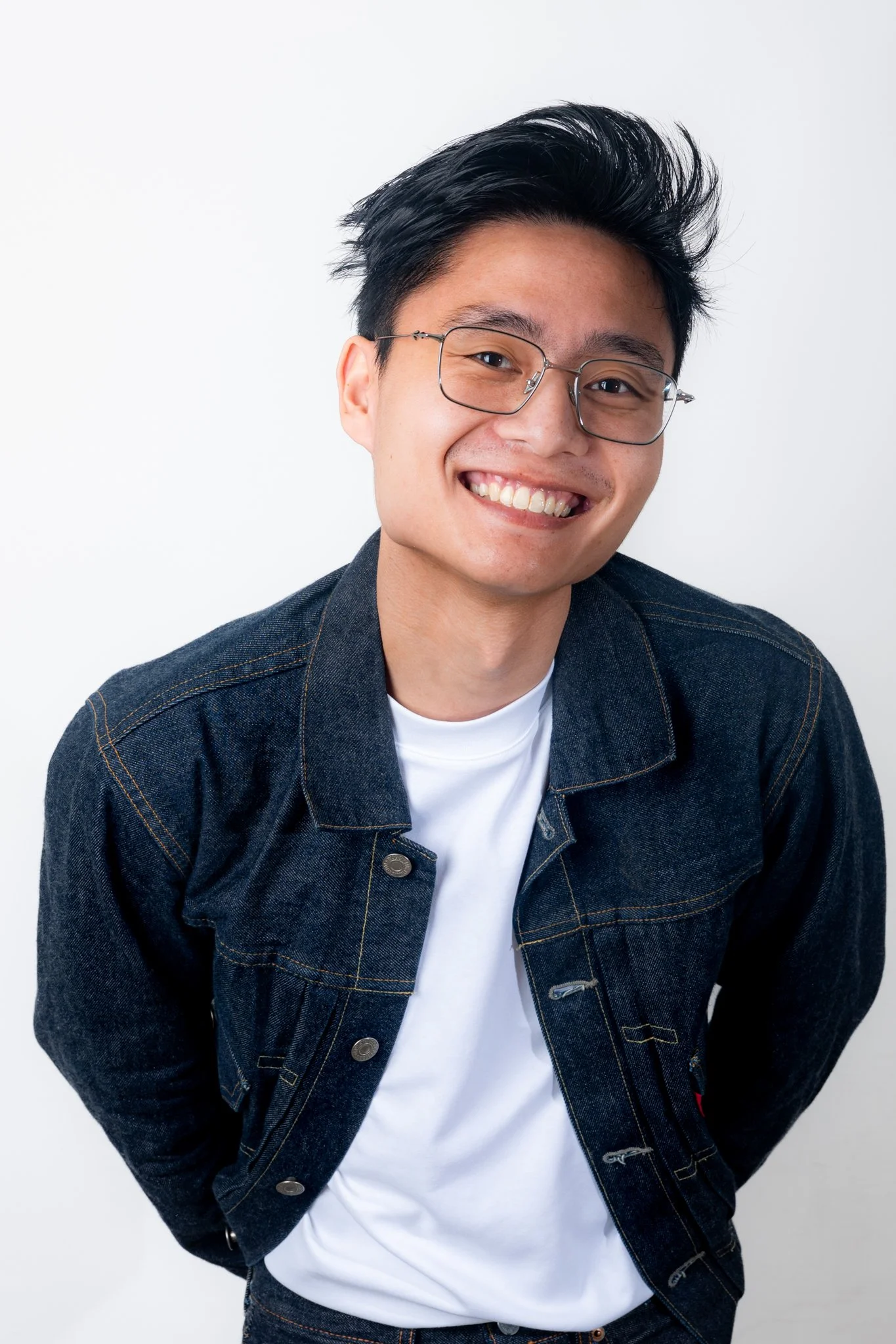 Smiling young man with glasses, wearing a denim jacket and white t-shirt, posing against a white background.