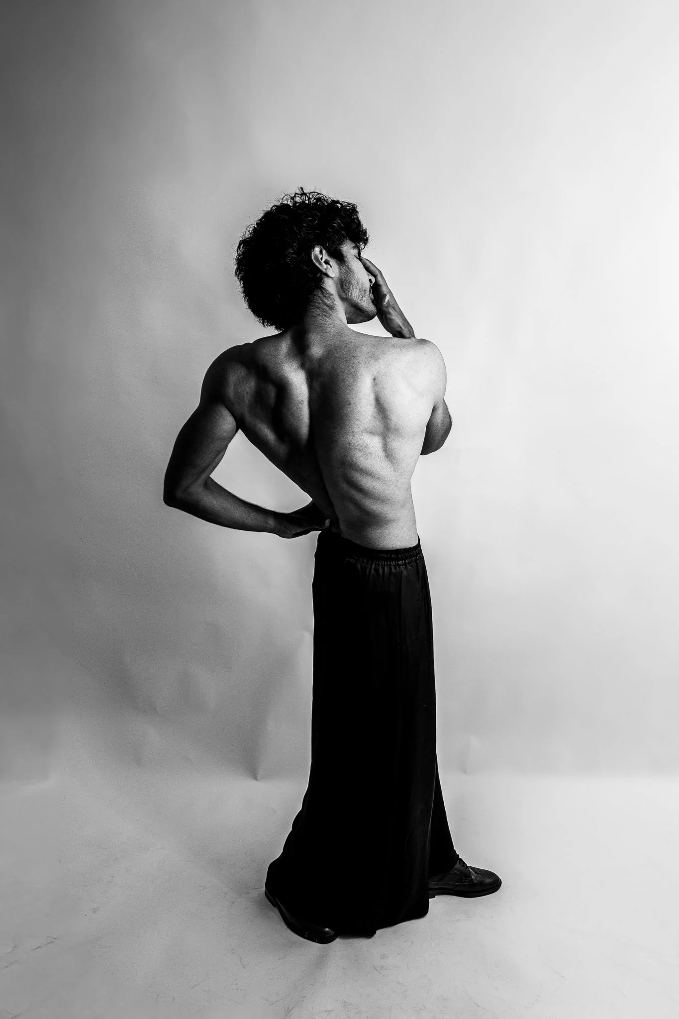 Black and white photo of a shirtless man standing sideways with one hand on his head and the other on his hip, in front of a plain background.