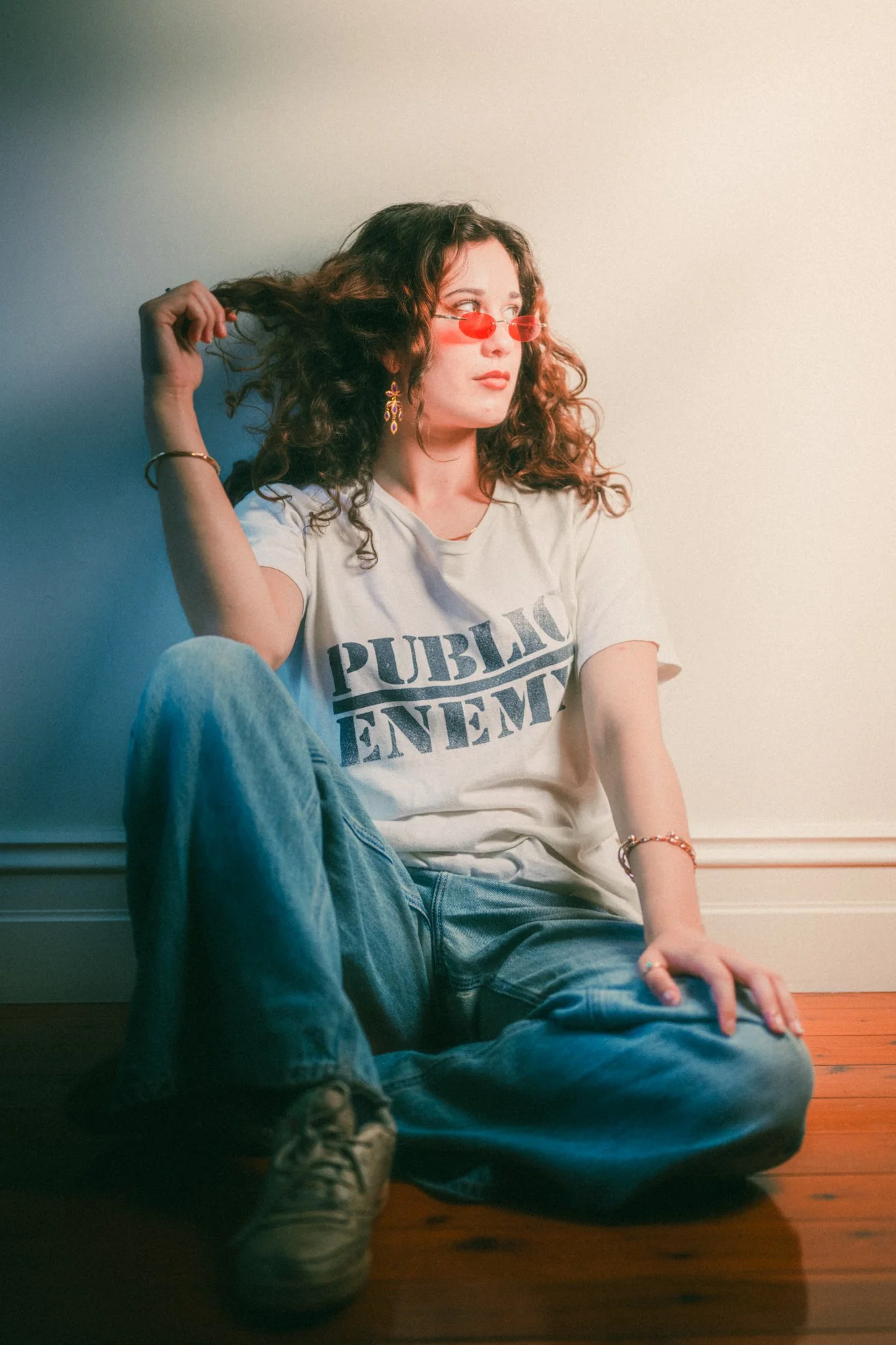 A woman with curly hair wearing red-tinted sunglasses, a white T-shirt with 'PUBLIC ENEMY' printed on it, blue jeans, and sneakers, sitting on the floor against a plain wall.