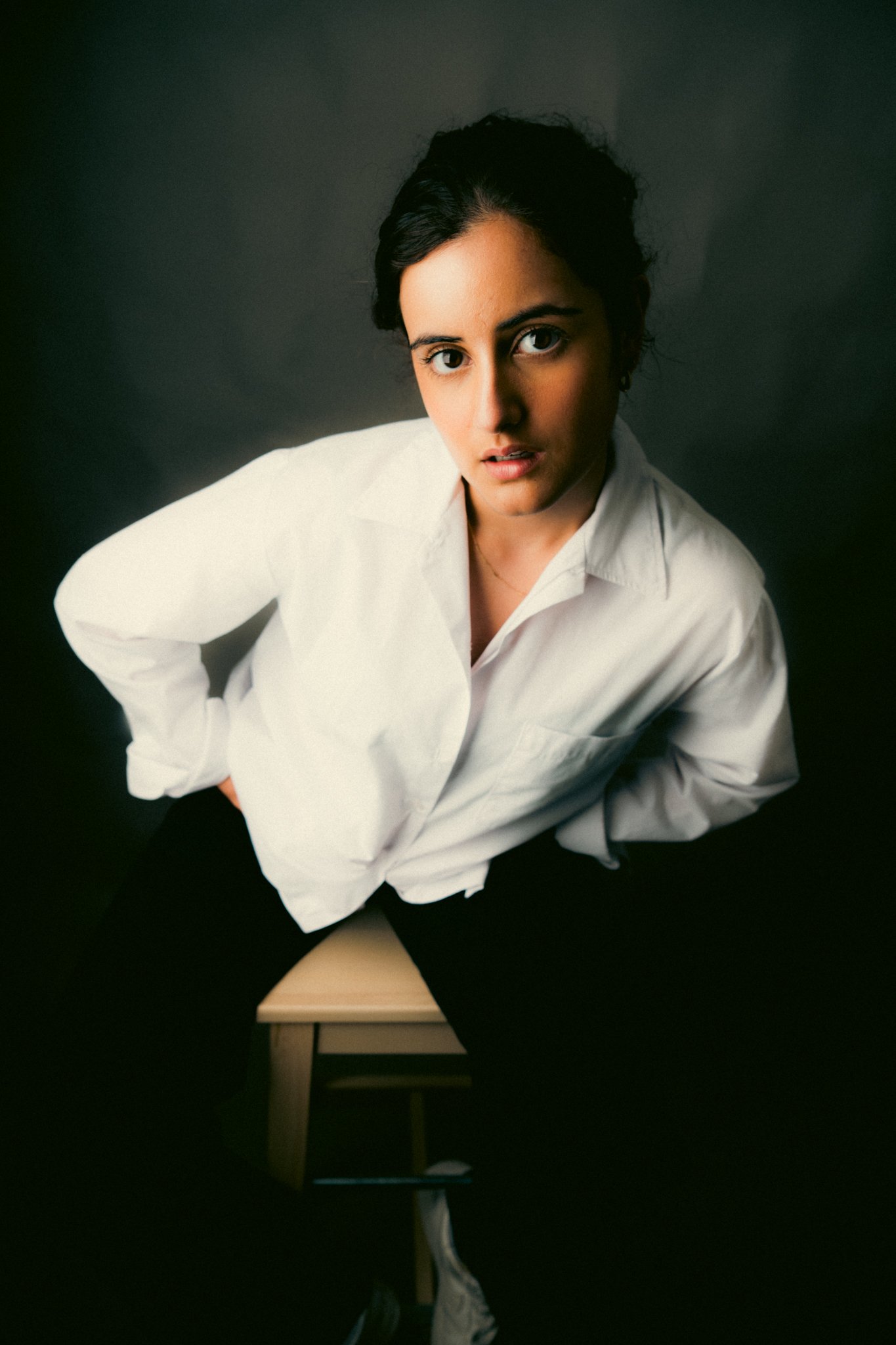 Portrait of a woman with dark hair wearing a white shirt, seated on a wooden stool with a dark background.