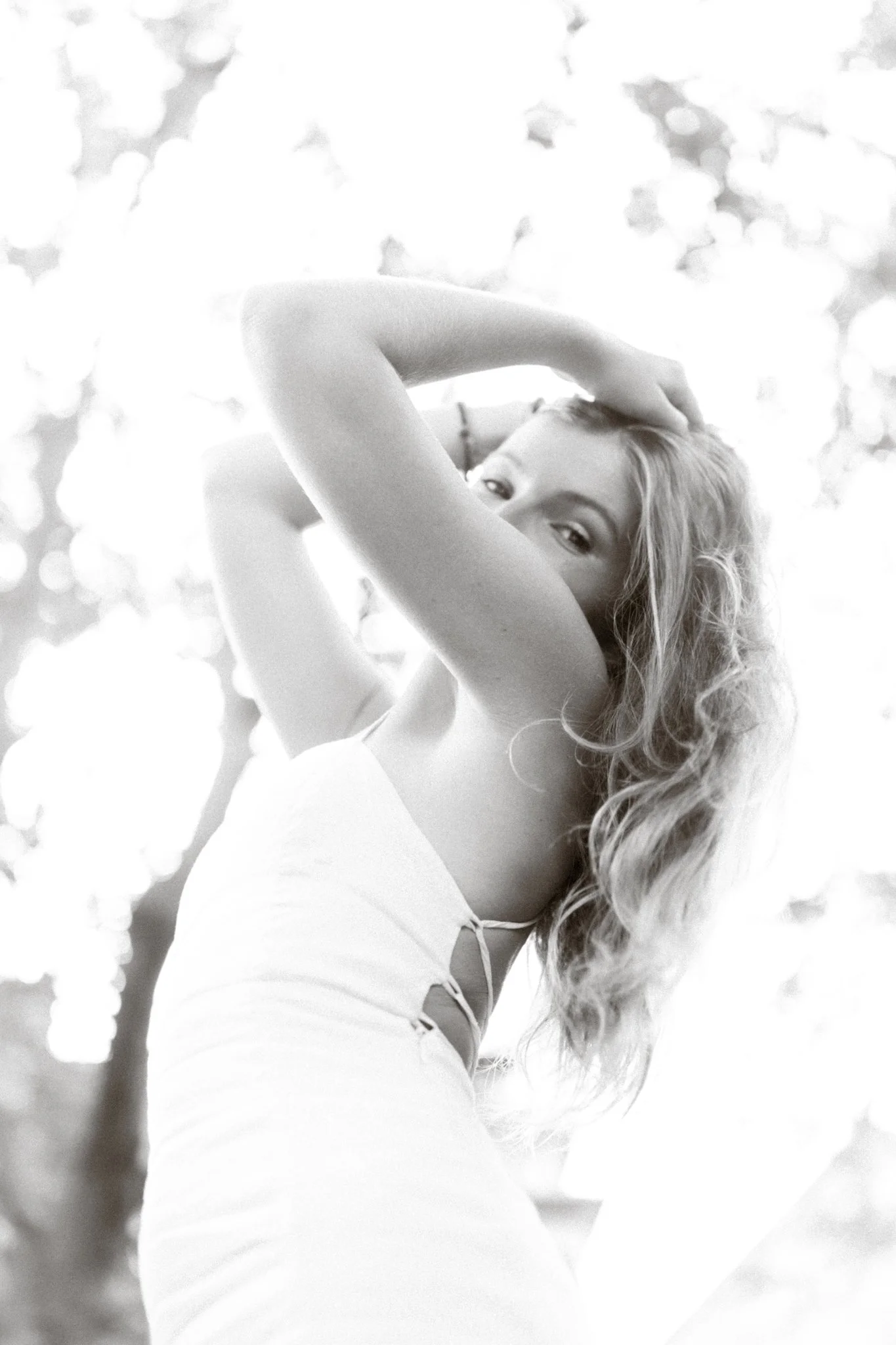 A woman with long, wavy hair looks over her shoulder while smiling, with her arms raised and hands on her head, standing outdoors.