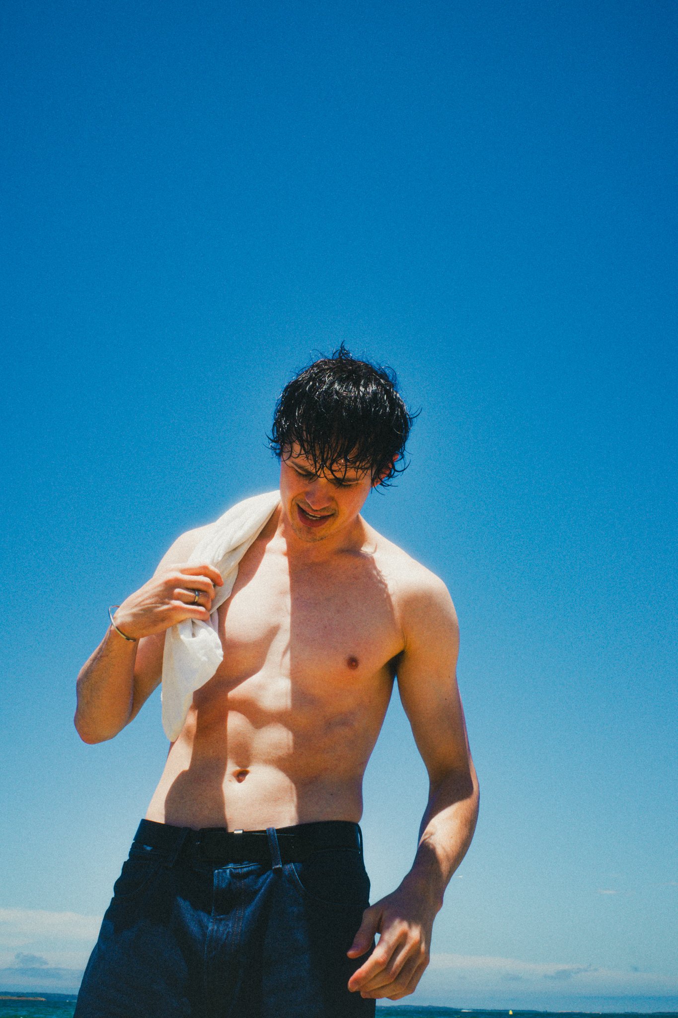 A shirtless man with wet hair holding a towel over his shoulder, standing outdoors under a clear blue sky.