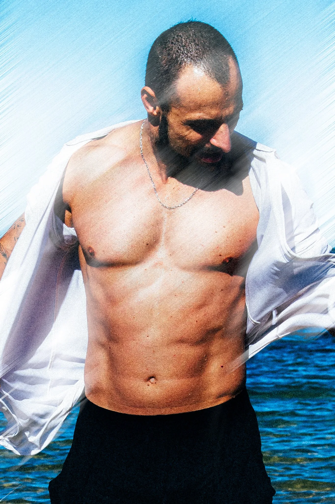 A shirtless man with a beard and short hair outdoors by the water, with a white jacket draped over his shoulders and a silver chain necklace, sunlight casting shadows.