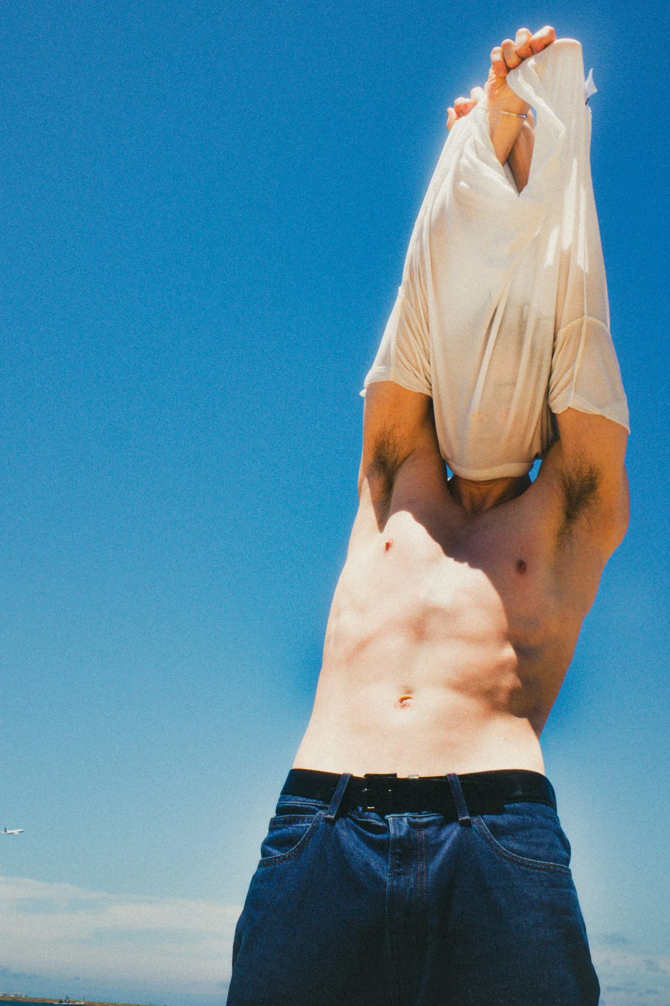 A man with exposed abs lifting a shirt over his head outdoors against a bright blue sky.