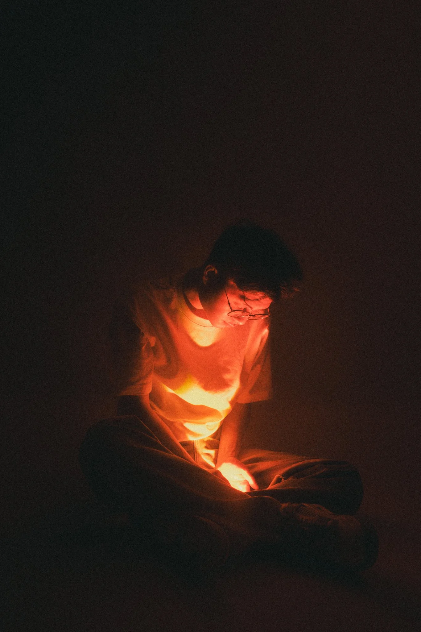 Person sitting cross-legged, illuminated by a warm, orange light source, in a dark environment.