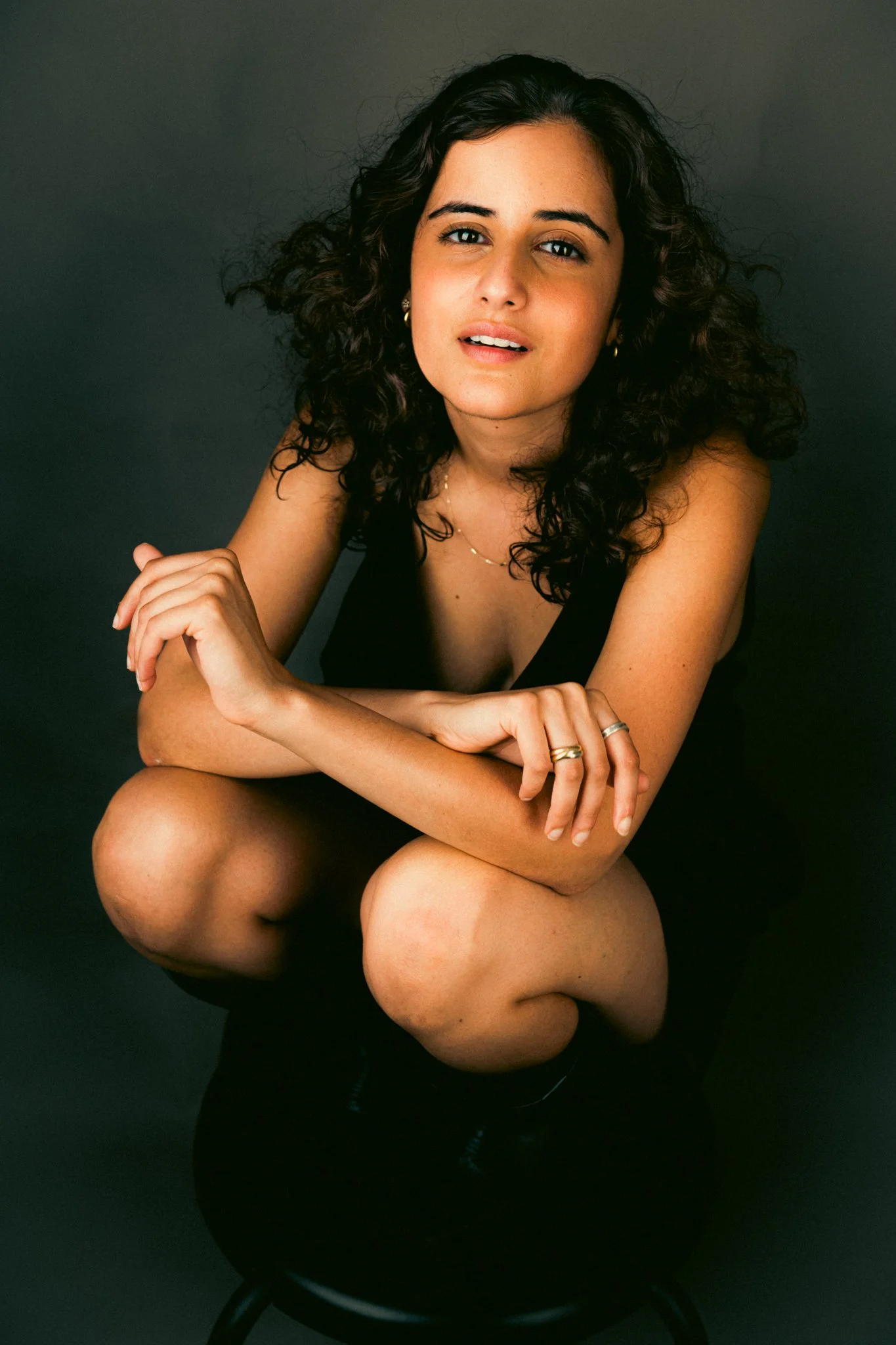 A woman with curly dark hair, wearing a black top, sitting on a black stool with her knees drawn up, against a dark background.