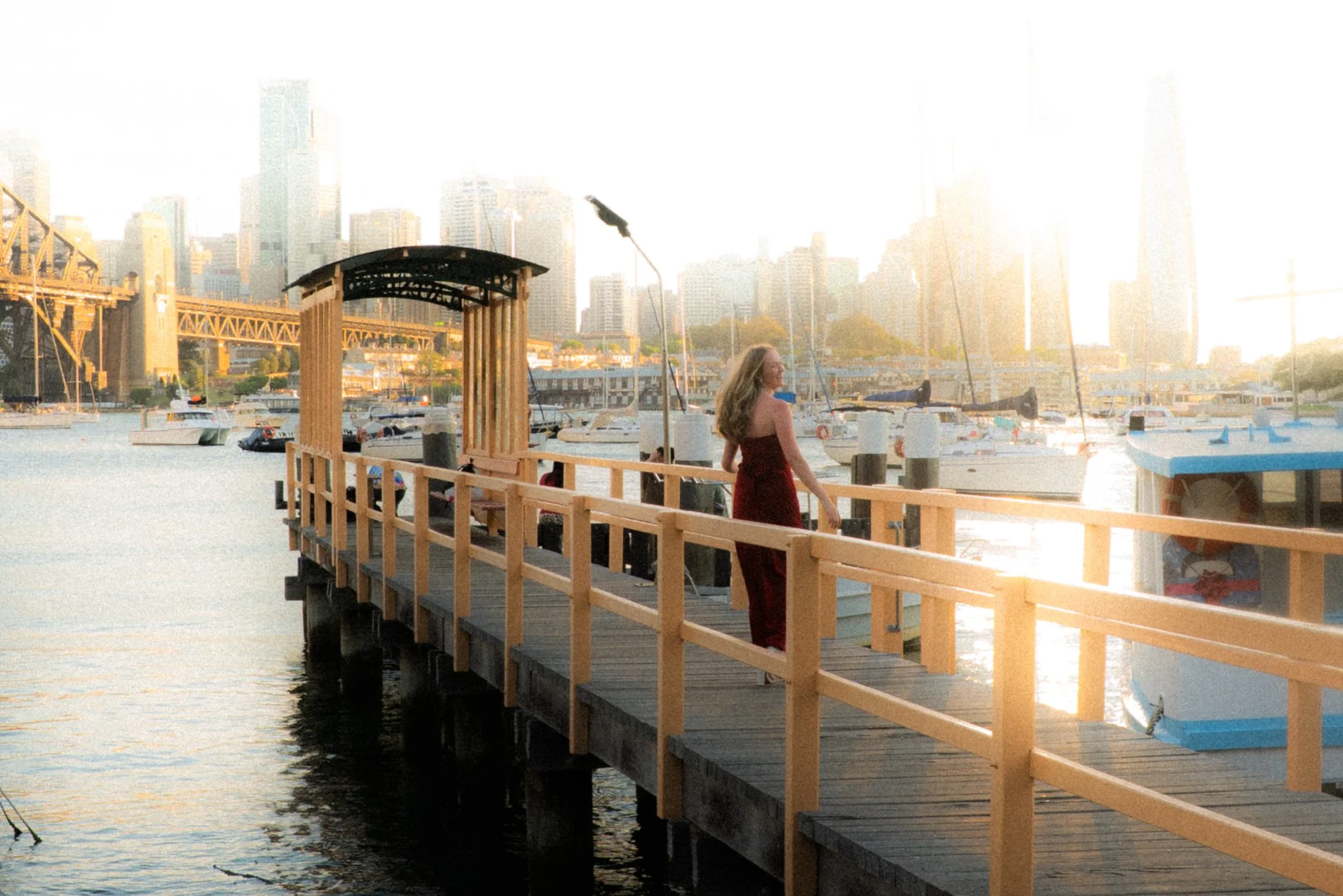 A woman in a red dress standing on a wooden pier by the water with boats and a city skyline in the background at sunset.