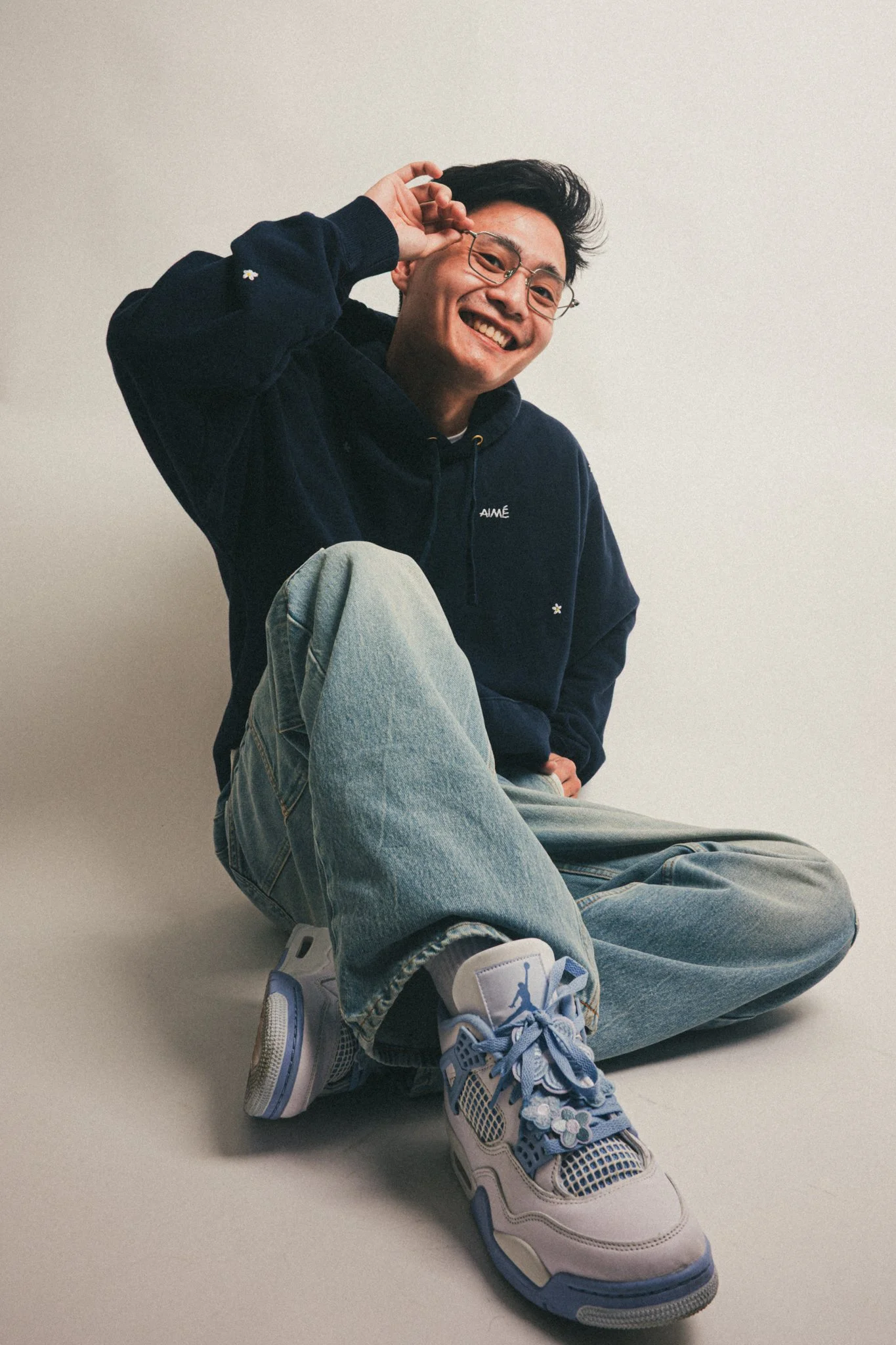 A young man sitting on the floor, smiling, wearing glasses, a dark hoodie, light blue jeans, and pastel-colored sneakers.